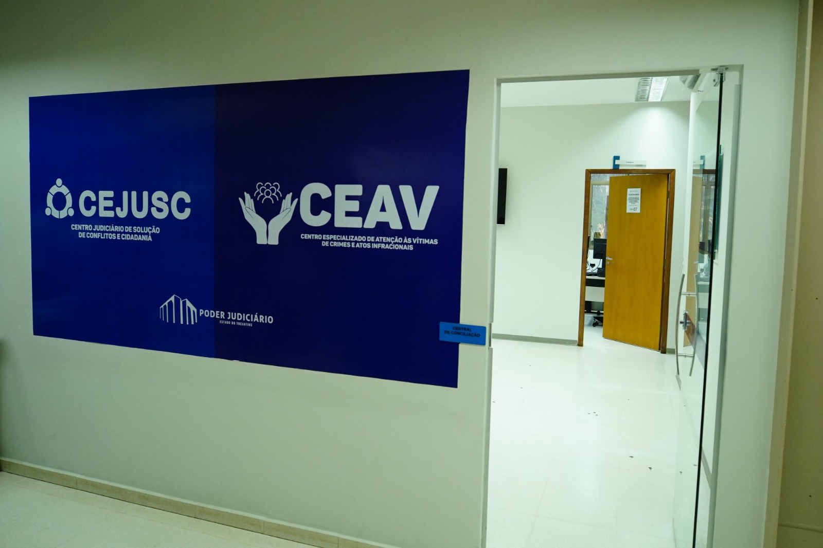 Fotografia colorida que mostra um painel azul com letras brancas escritas Cejusc e Ceav, o painel está na entrada de uma sala