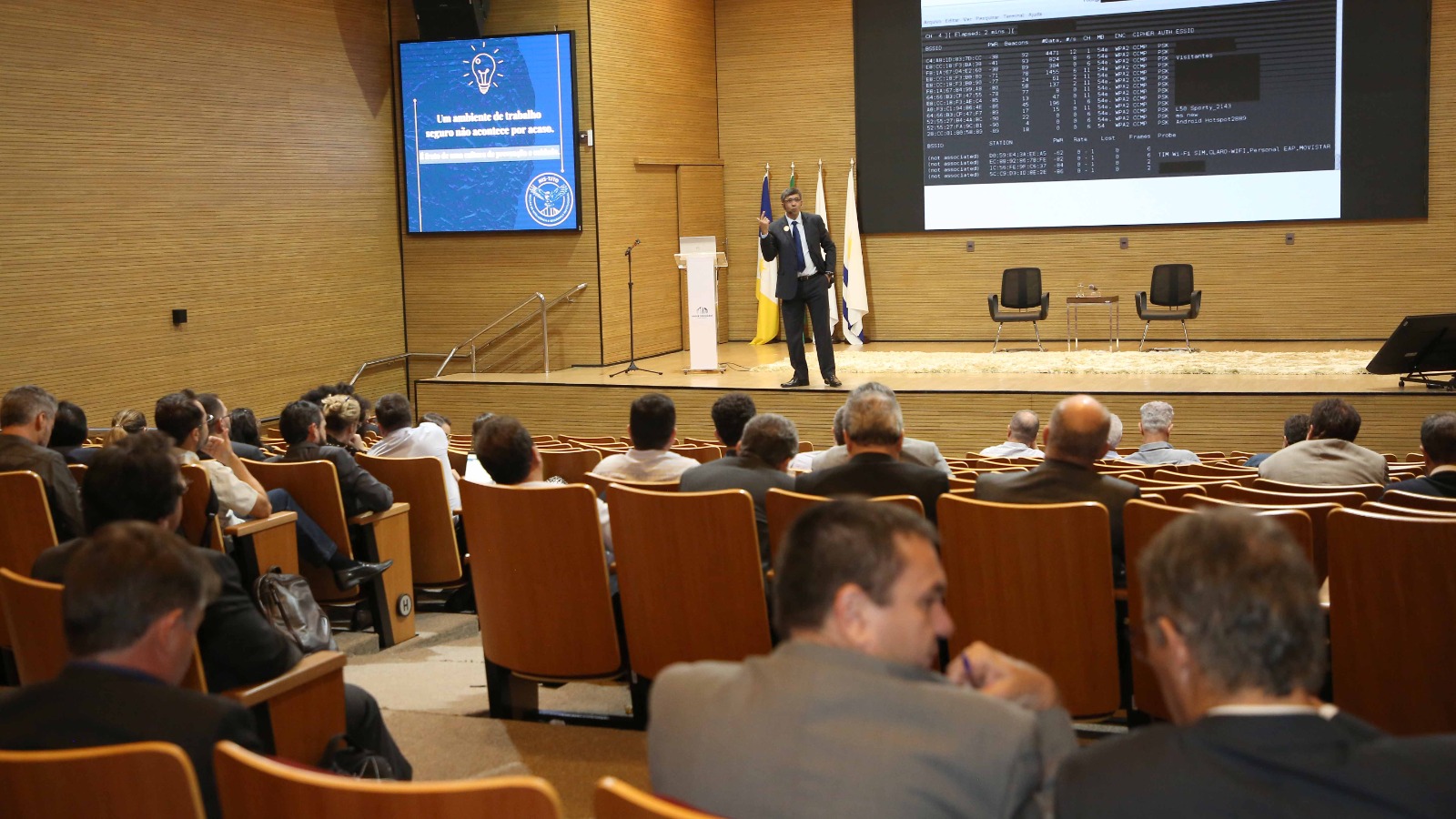 Homem moreno e cabelos curtos lisos, de terno escuro, está em cima do palco do auditório do TJTO dando palestra para plateia que está de costas formada por homens e mulheres 