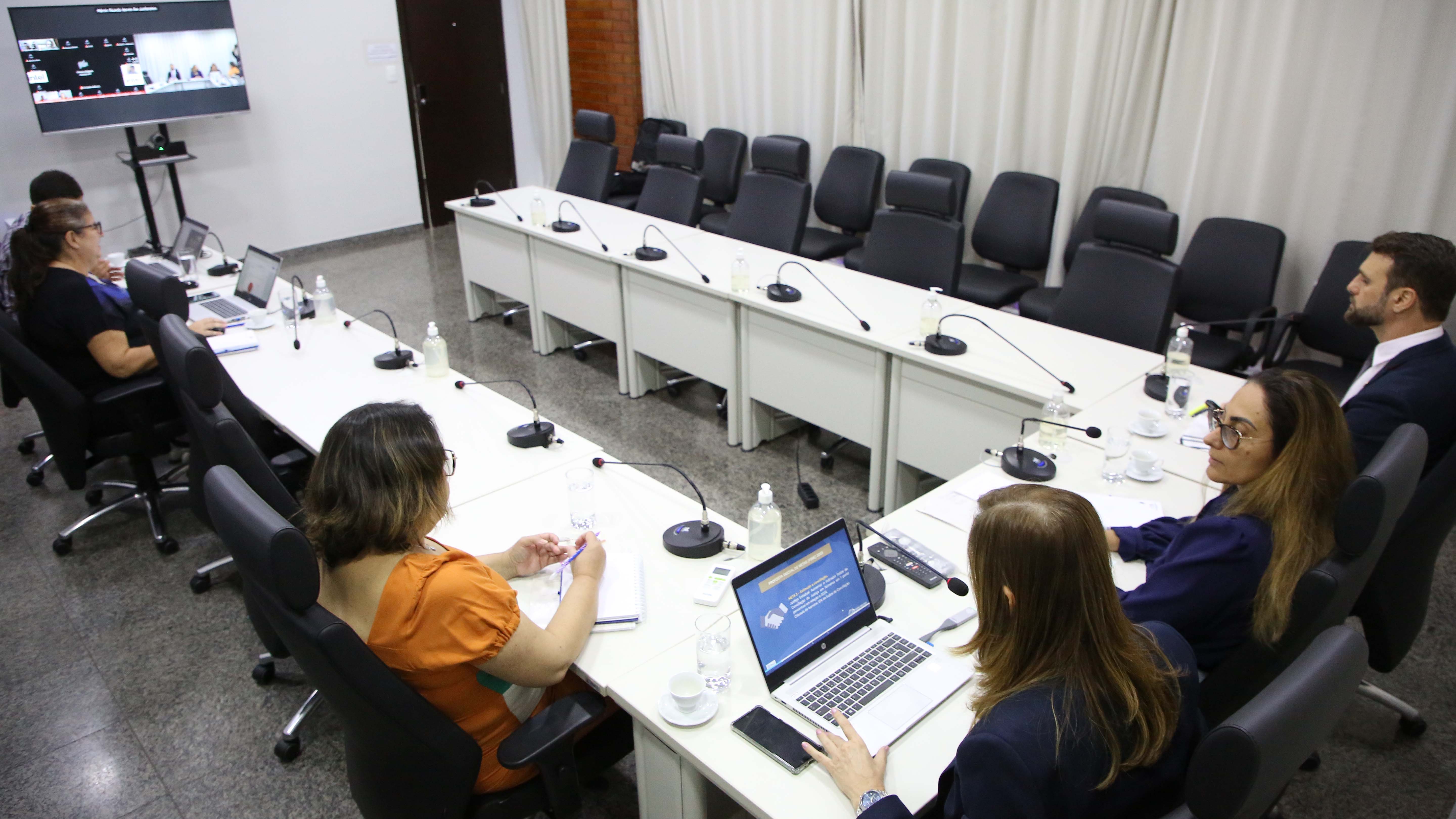 Homens e mulheres sentados em torno de mesa branca em formato U, com microfones individuais, durante reunião do TJTO sobre metas do CNJ, numa sala de paredes brancas com cortina branca