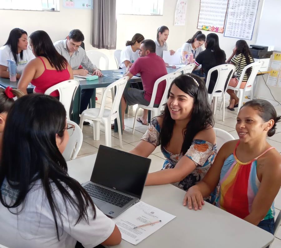 Imagem colorida que mostra duas mulheres sendo atendidas por outra mulher em uma sala com outras pessoas (homens e mulheres sendo atendidos)