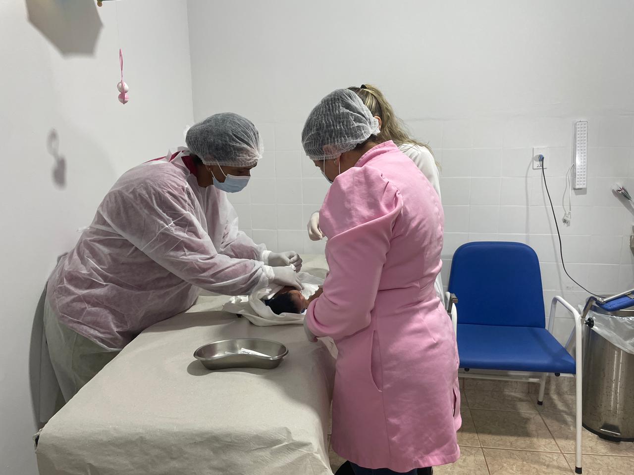 Três mulheres com jaleco vacinam um bebê que está em uma maca