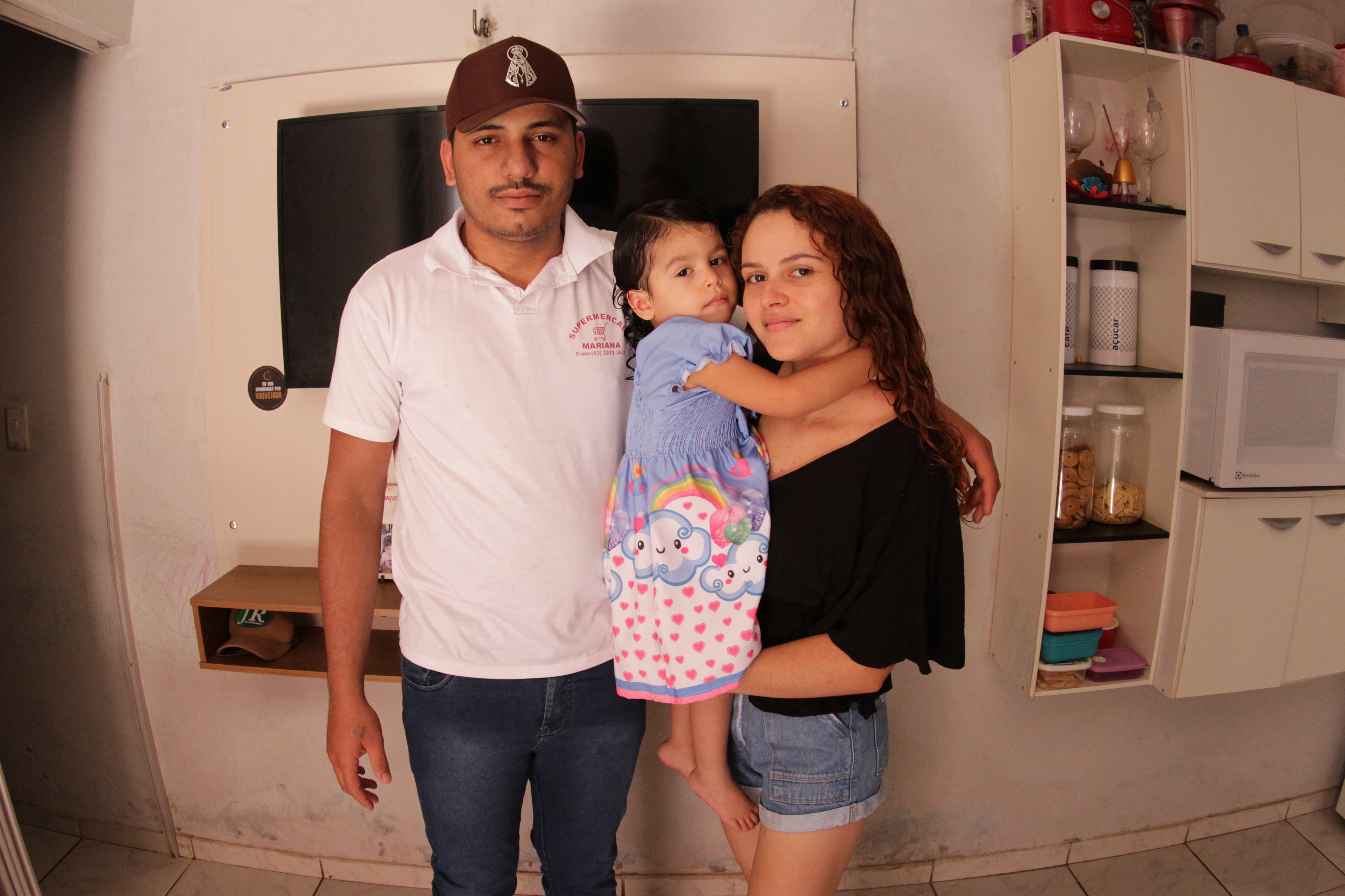 Fotografia colorida que mostra um homem de boné marrom e camisa rosa, com uma menina no colo que usa vestido lilás e ao lado de uma mulher ruiva de cabelos longos e encaracolados. 