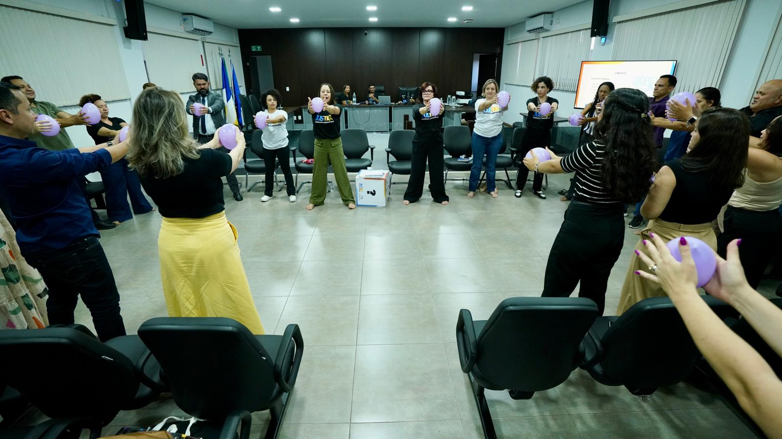 Pessoas em pé seguram balão com cor lilás e fazem exercícios de ginástica laboral