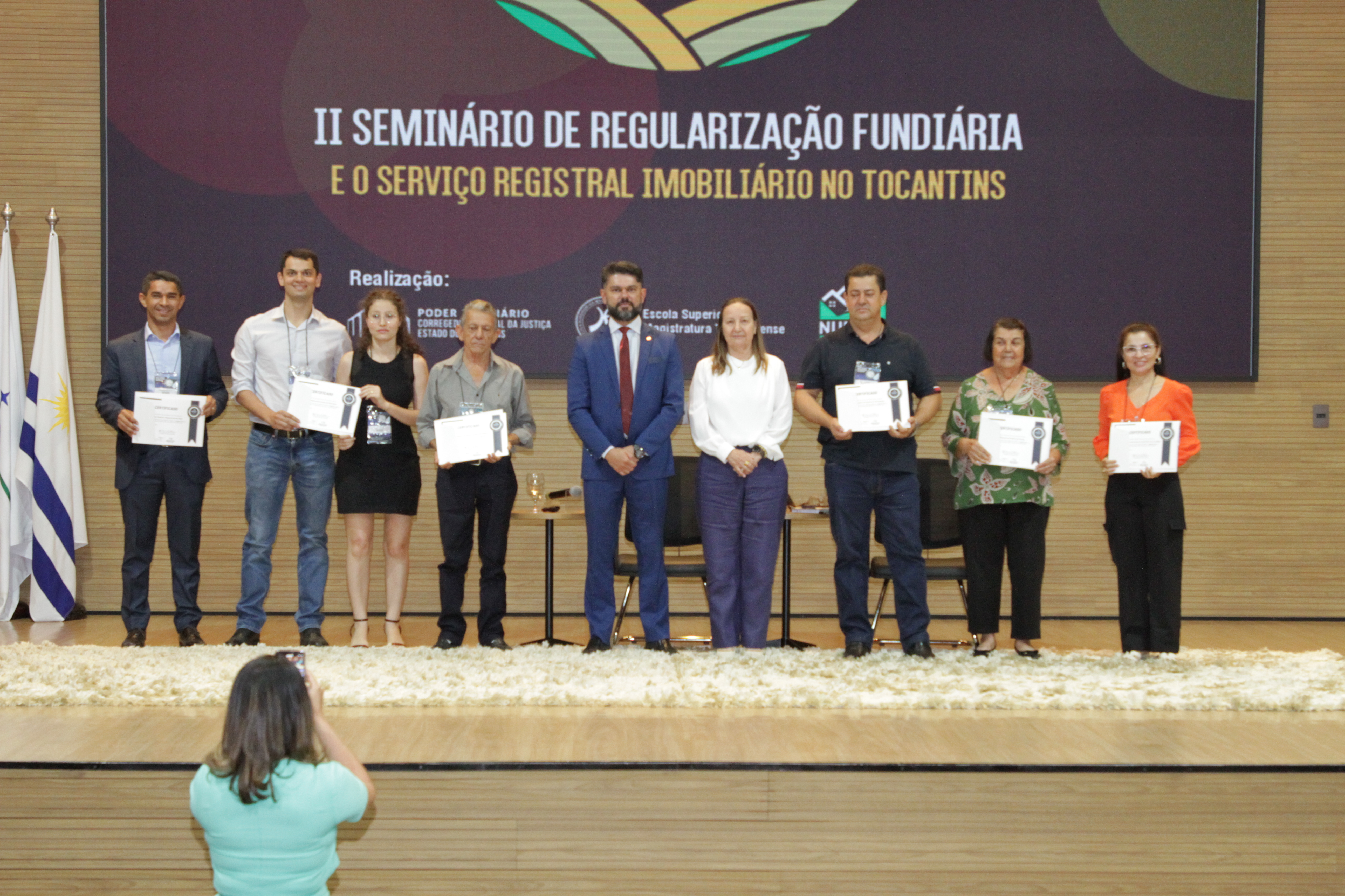 Fotografia colorida que mostra pessoas no palco segurando um certificado