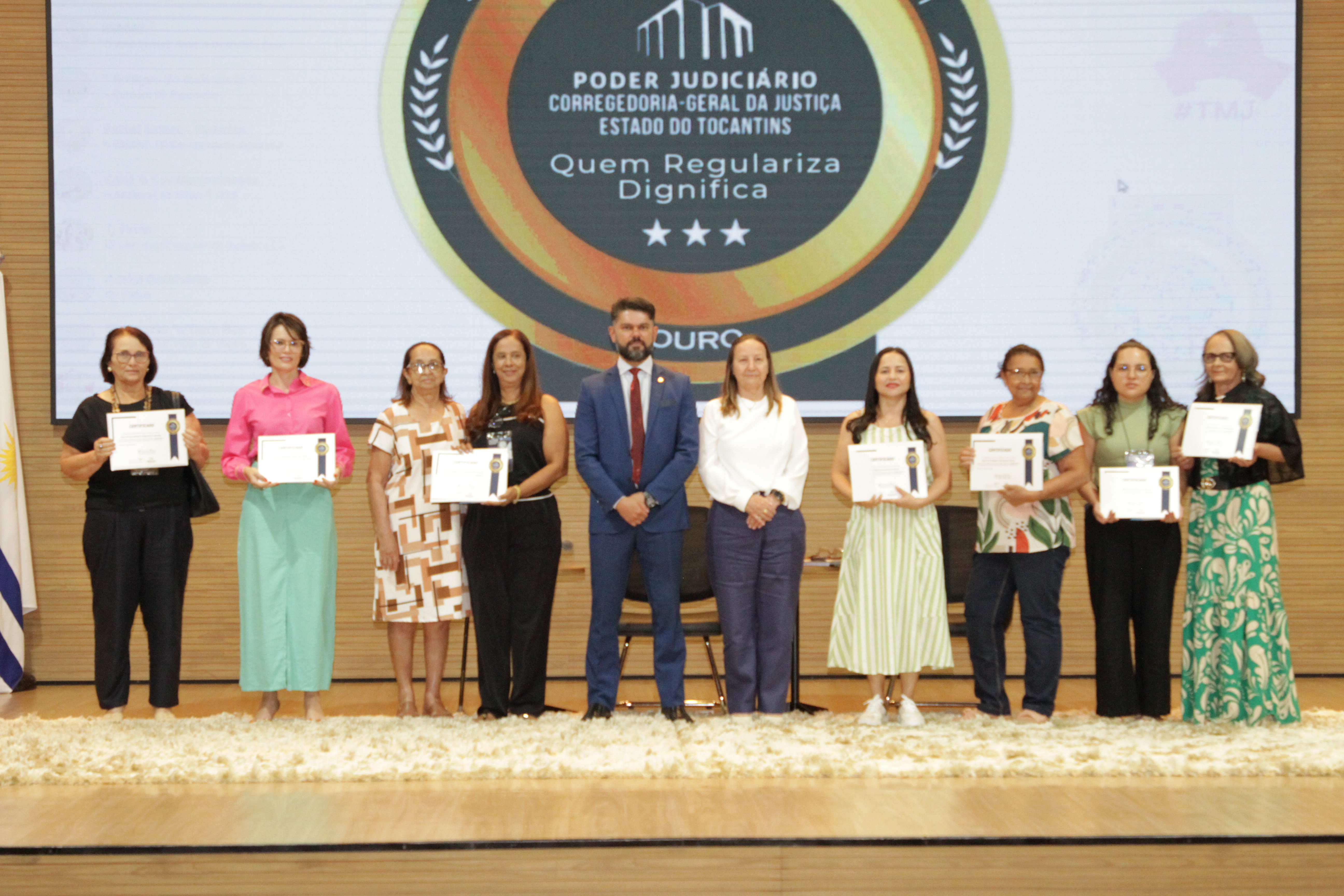 Fotografia colorida que mostra pessoas no palco segurando um diploma