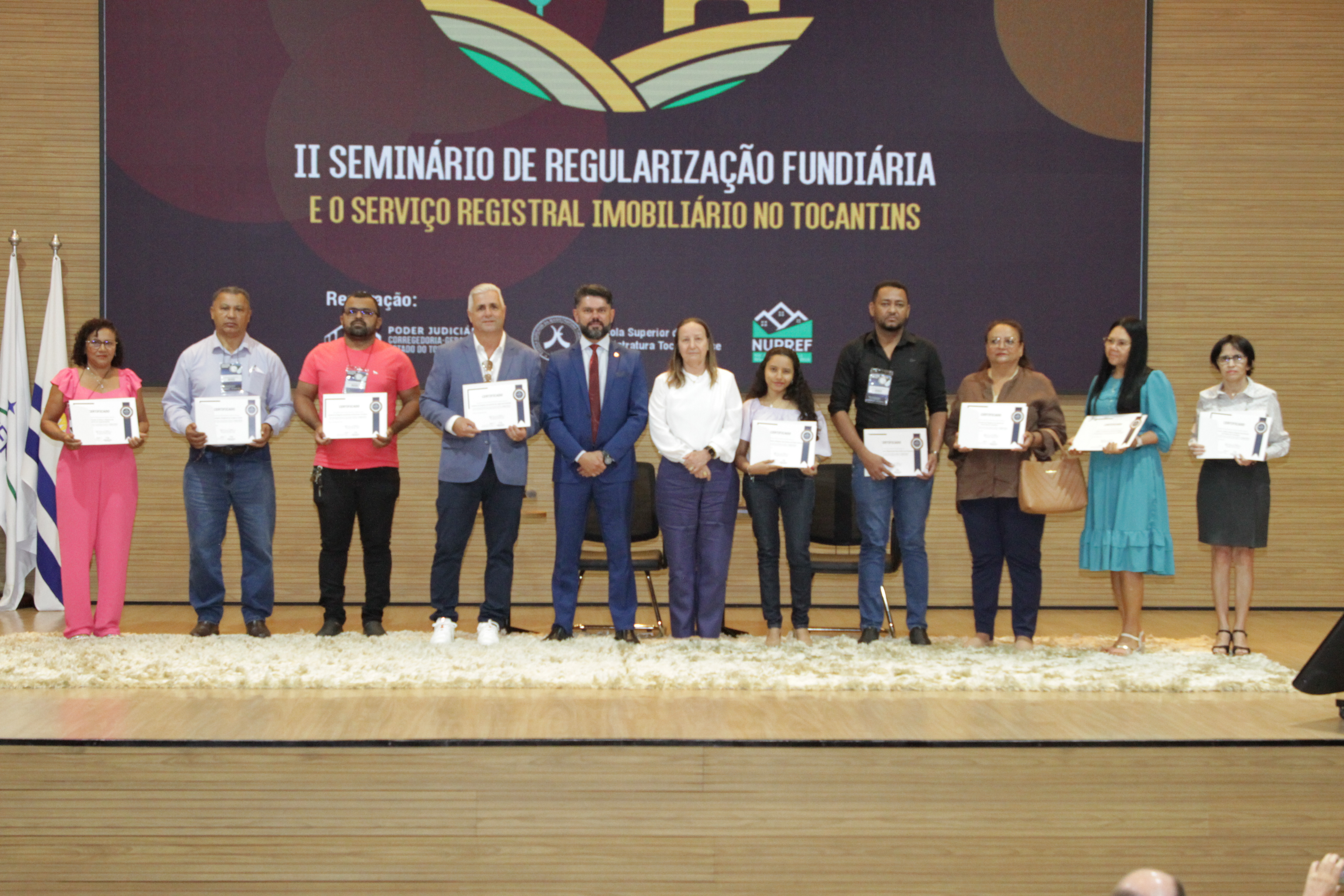 Fotografia colorida que mostram pessoas no palco segurando um certificado