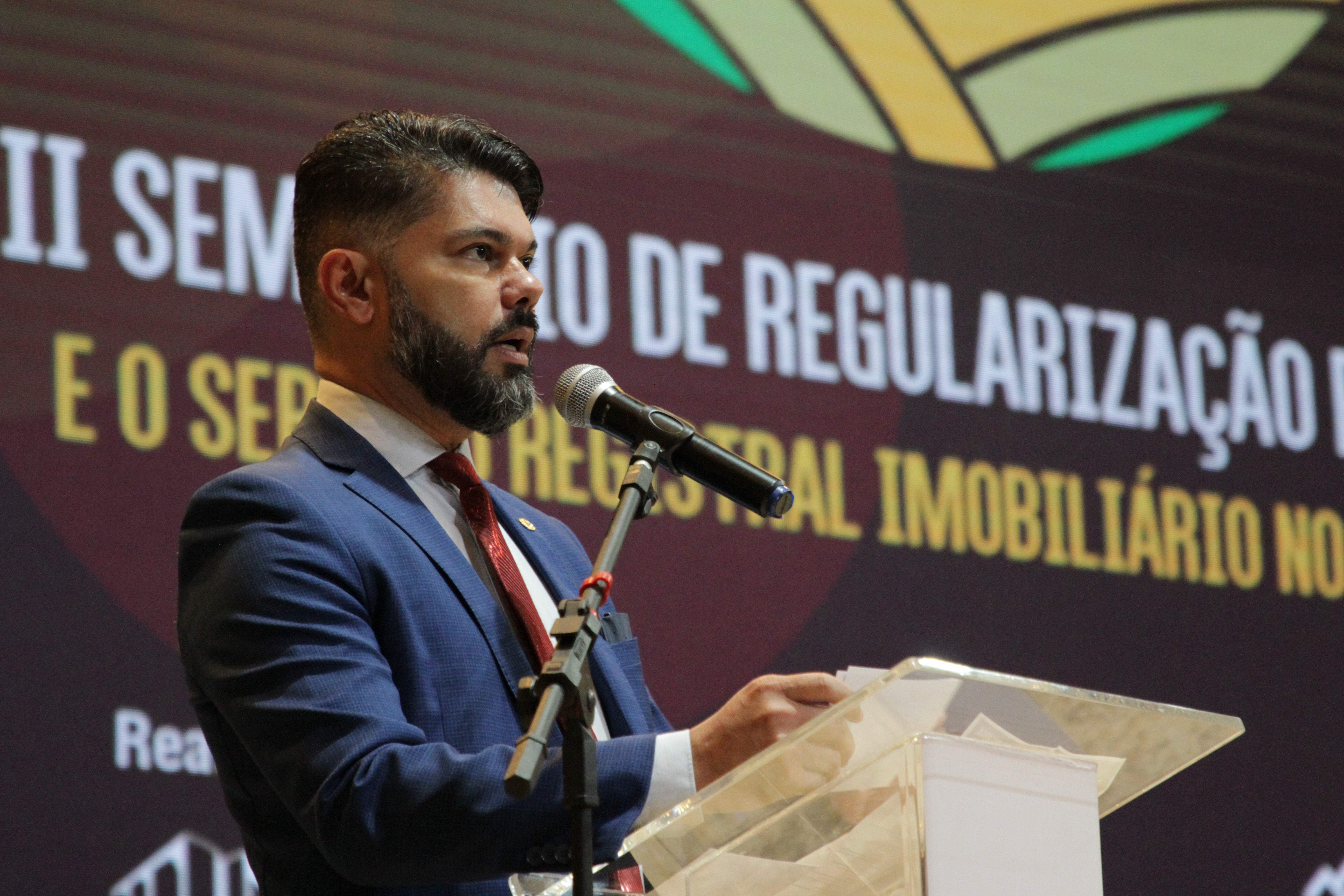 Imagem colorida que mostra um homem moreno de barba, que traja terno, e está falando ao microfone