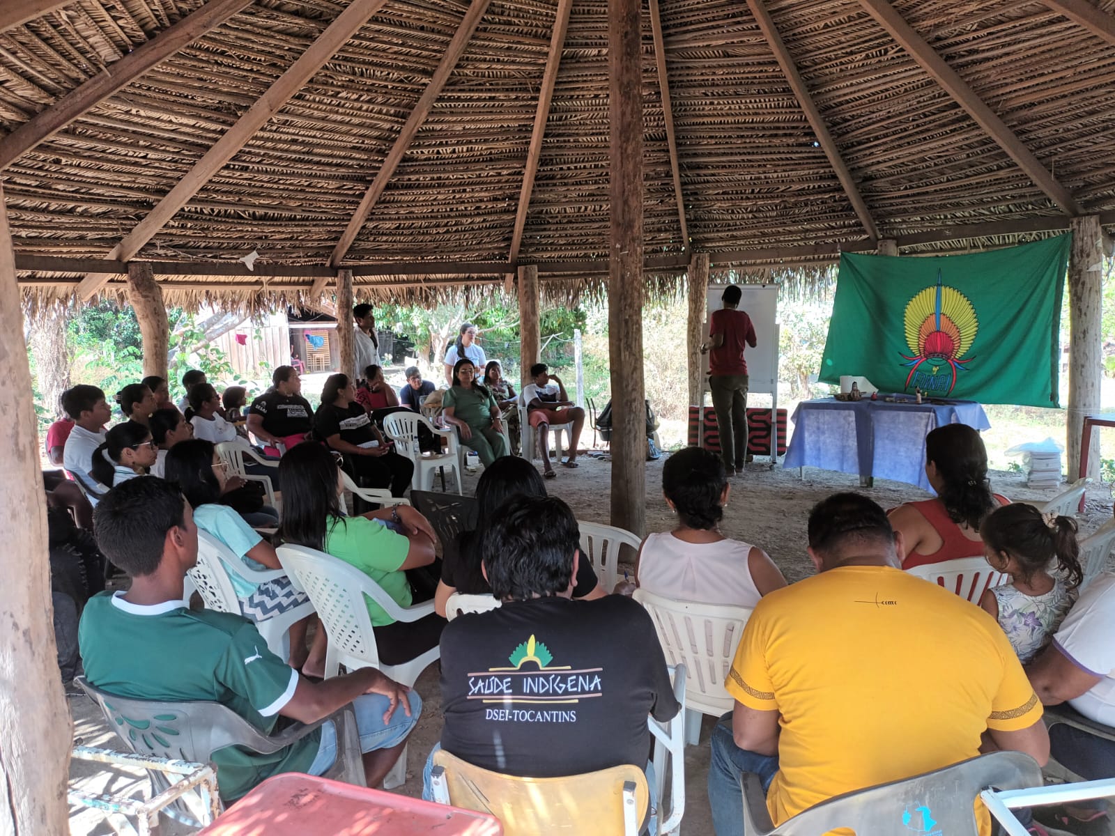 Indígenas reunidos embaixo de uma estrutura de palha assistindo a uma palestra