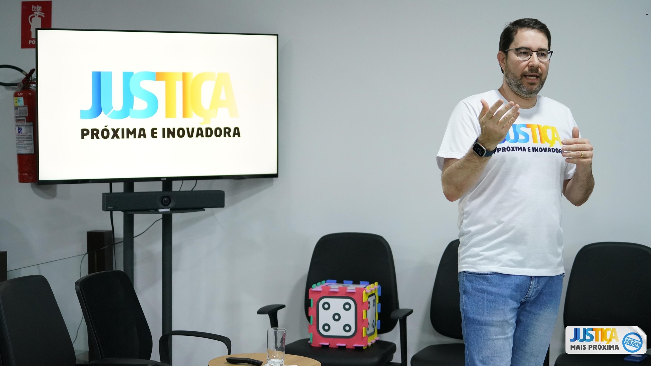 Fotografia colorida que mostra um homem branco usando jeans e camiseta escrita Justiça próxima e inovadora falando a uma plateia e do lado dele há uma TV com a logomarca Justiça mais proxima e inovadora