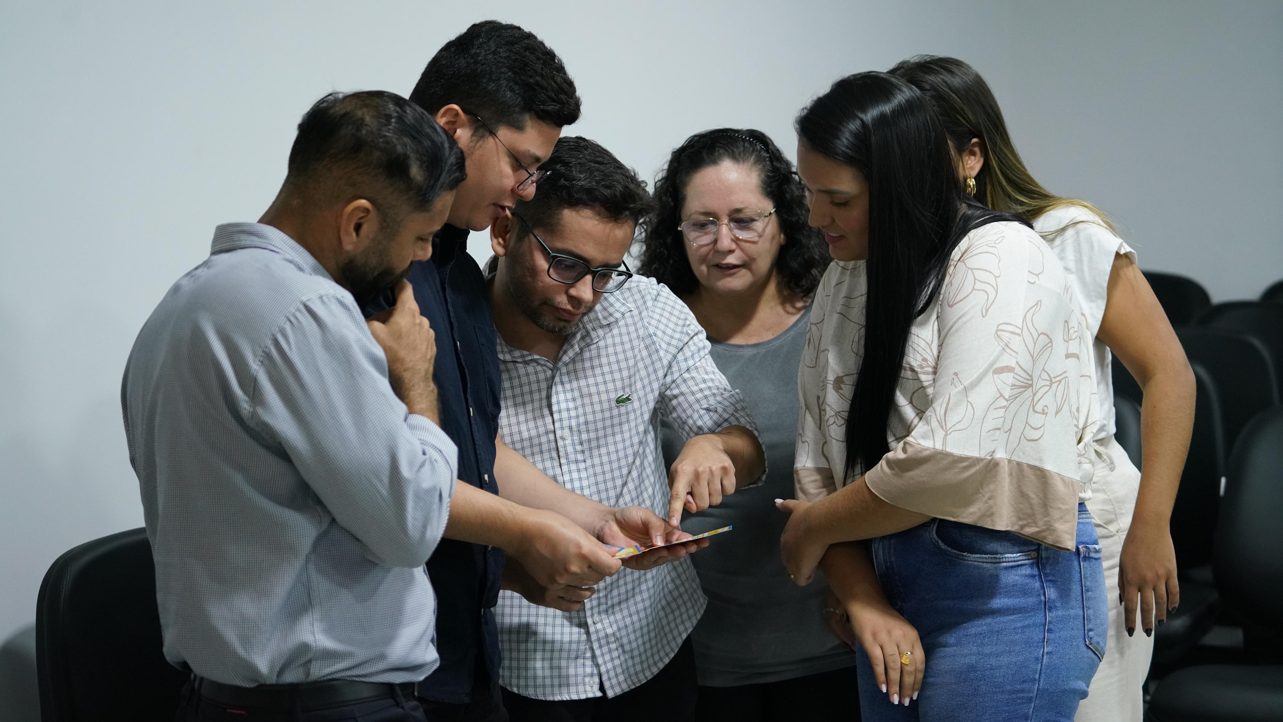 Imagem colorida que mostra homens e mulheres segurando cartas e observando