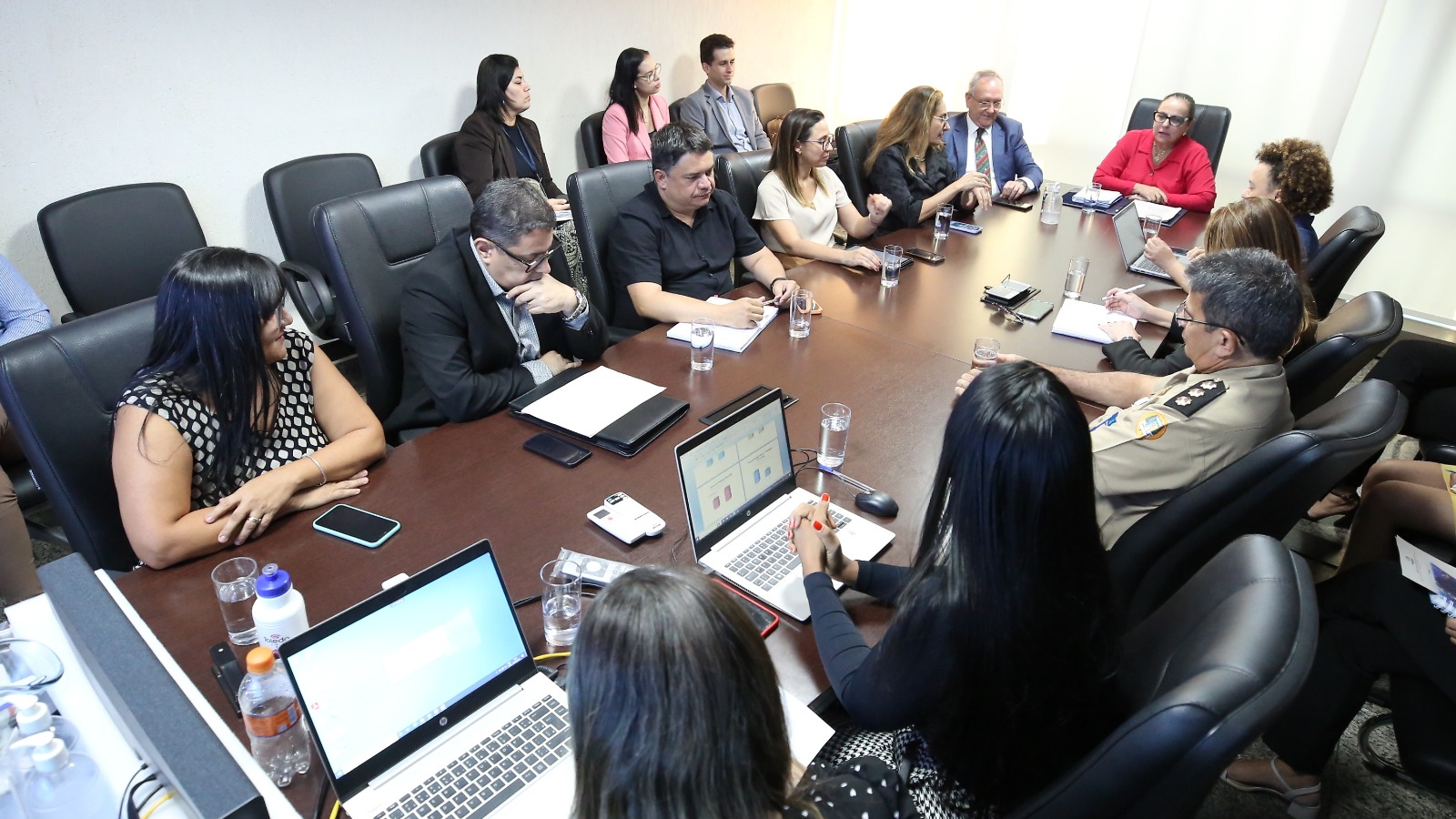 Homens e mulheres reunidos sentados em cadeiras pretas em torno de mesa de madeira numa sala de paredes brancas