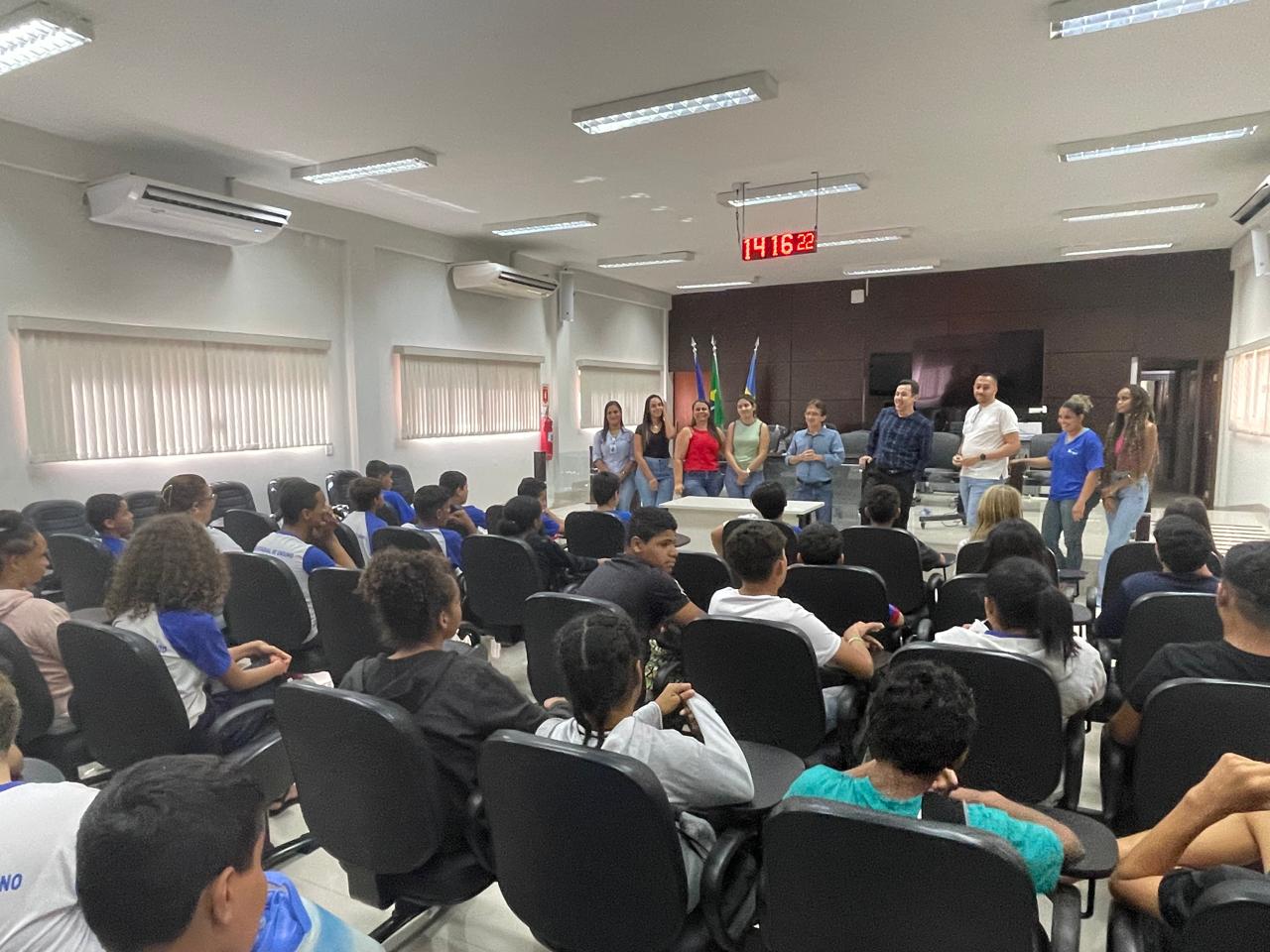 Crianças sentadas de costas no Salão do Júri. Á frente, homens e mulheres em pé falam sobre a carreira na magistratura. A sala tem paredes brancas com persianas claras e verticais, ao fundo, um painel de madeira escuro, com mastro de bandeiras à esquerda