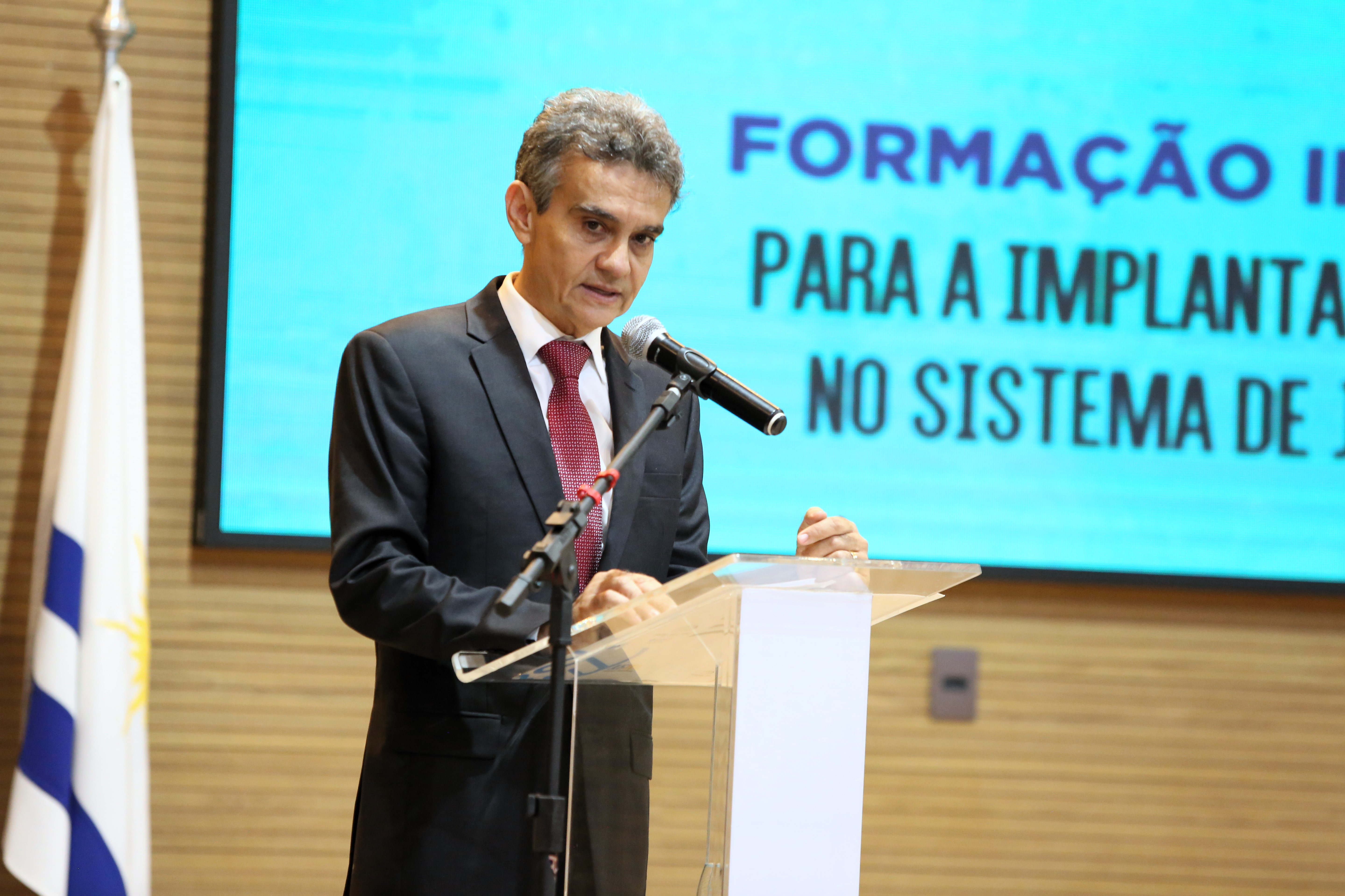 Fotografia colorida que mostra um homem branco, magro, usando terno marrom e gravata vermelha, falando ao microfone 