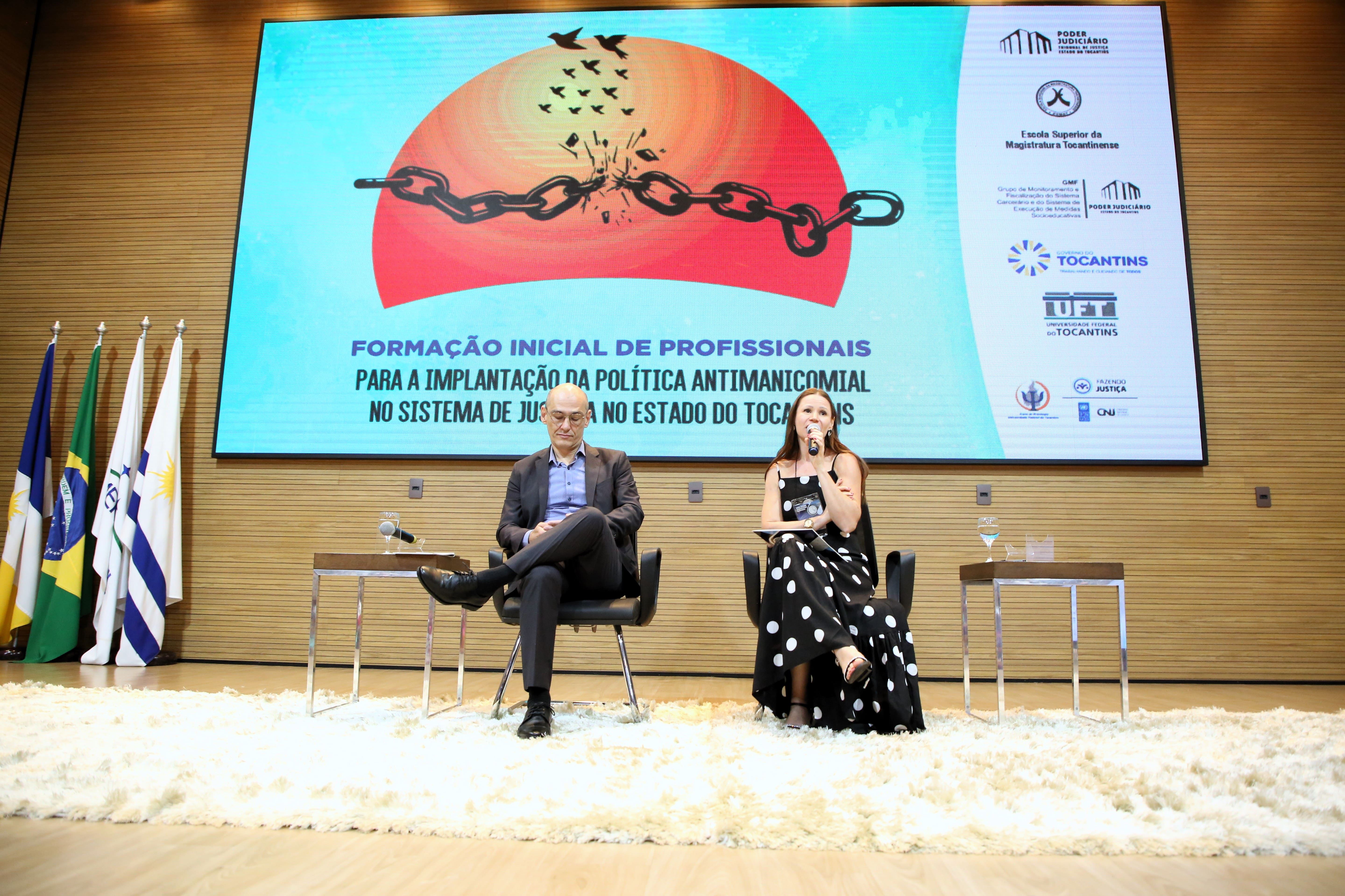 Fotografia colorida que mostra duas pessoas sentadas à frente de um palco, proferindo palestra