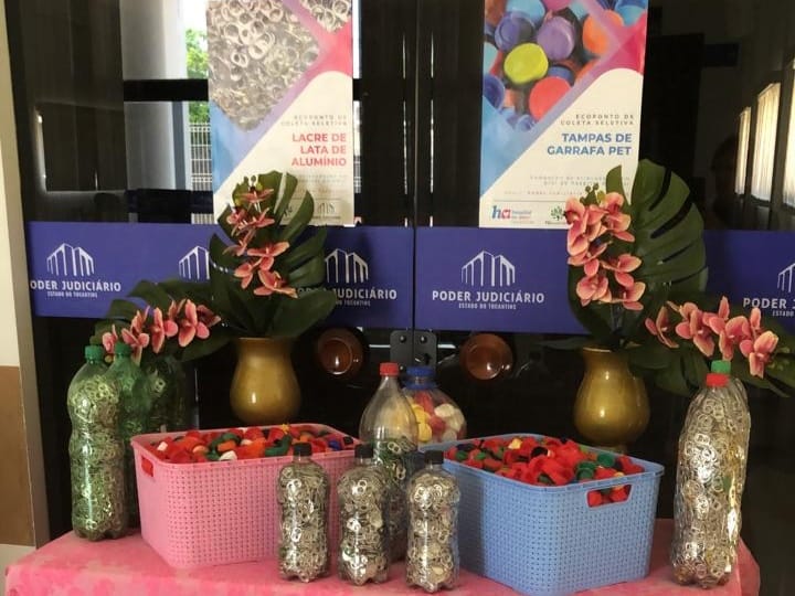 Porta de vidro com adesivo azul do Tribunal de Justiça do Tocantins e encartes de campanha de arrecadação de lacres e tampinhas para o Hospital de Amor. Na frente, mesa com toalha rosa e vasilhas com lacres e tampinhas em cima
