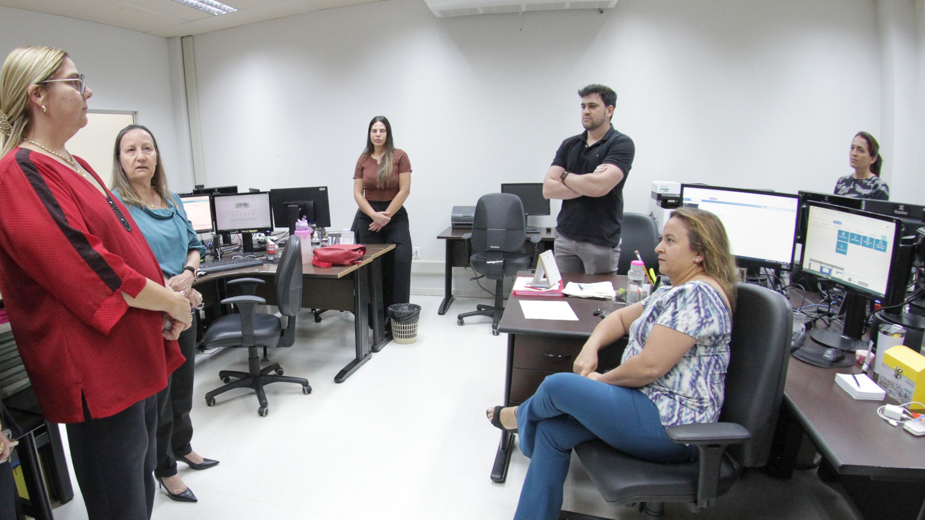 Fotografia colorida que mostra uma mulher loira usando vermelho e preto em pé, uma mulher branca de óculos usando azul e preto, também em pé, ambas falam para quatro pessoas que estão numa sala com computadores