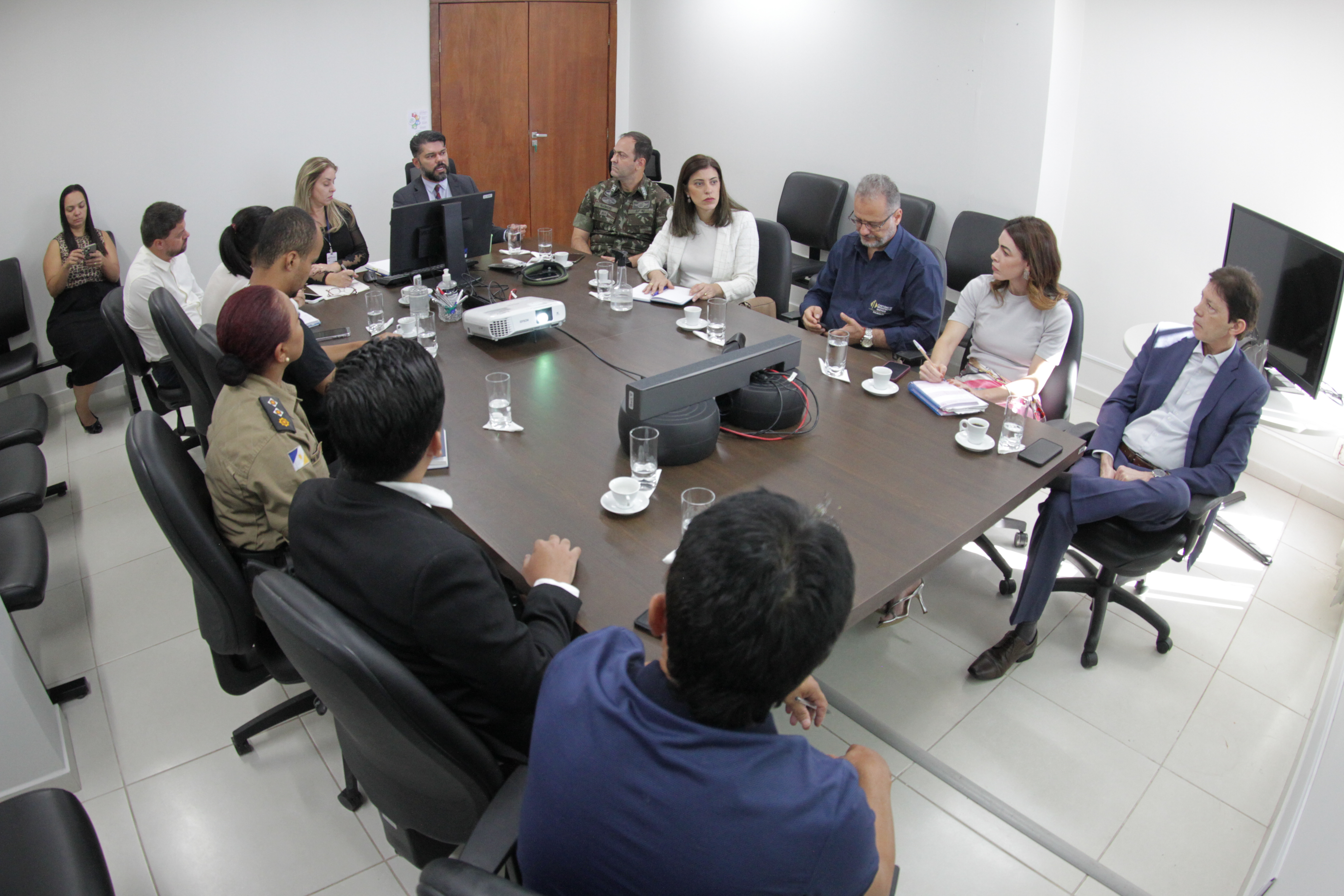 Fotografia colorida onde um grupo de treze pessoas estão sentados em volta de uma mesa retangular de madeira escura conversando. Dessas oito pessoas, oito são homens e cinco mulheres. Ao fundo, uma outra mulher está sentada em uma cadeira fora da mesa. Eles estão em uma sala branca.