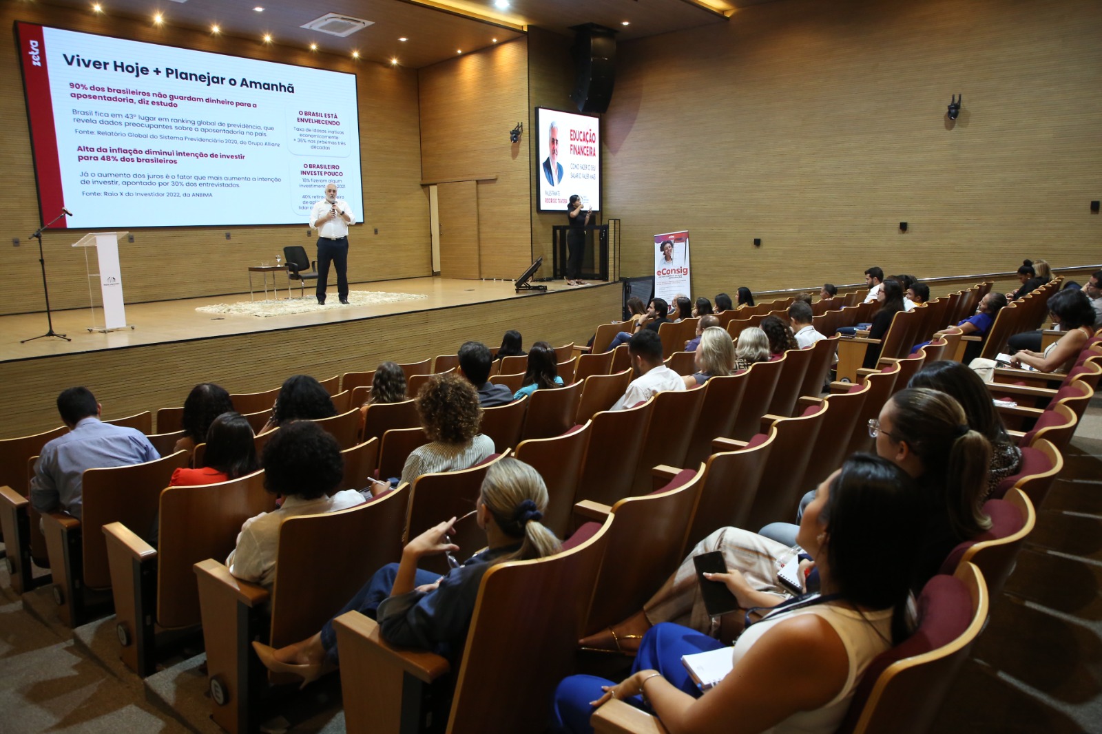 Imagem aberta do auditório do TJTO com vários servidores entados assistindo a palestra sobre educação financeira; ao fundo, no palco, o palestrante falando ao microfone e uma imagem projetada ao fundo. 