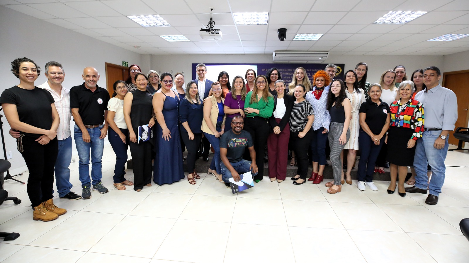 Fotografia colorida de muitas pessoas em pé, numa sala, posando para a foto