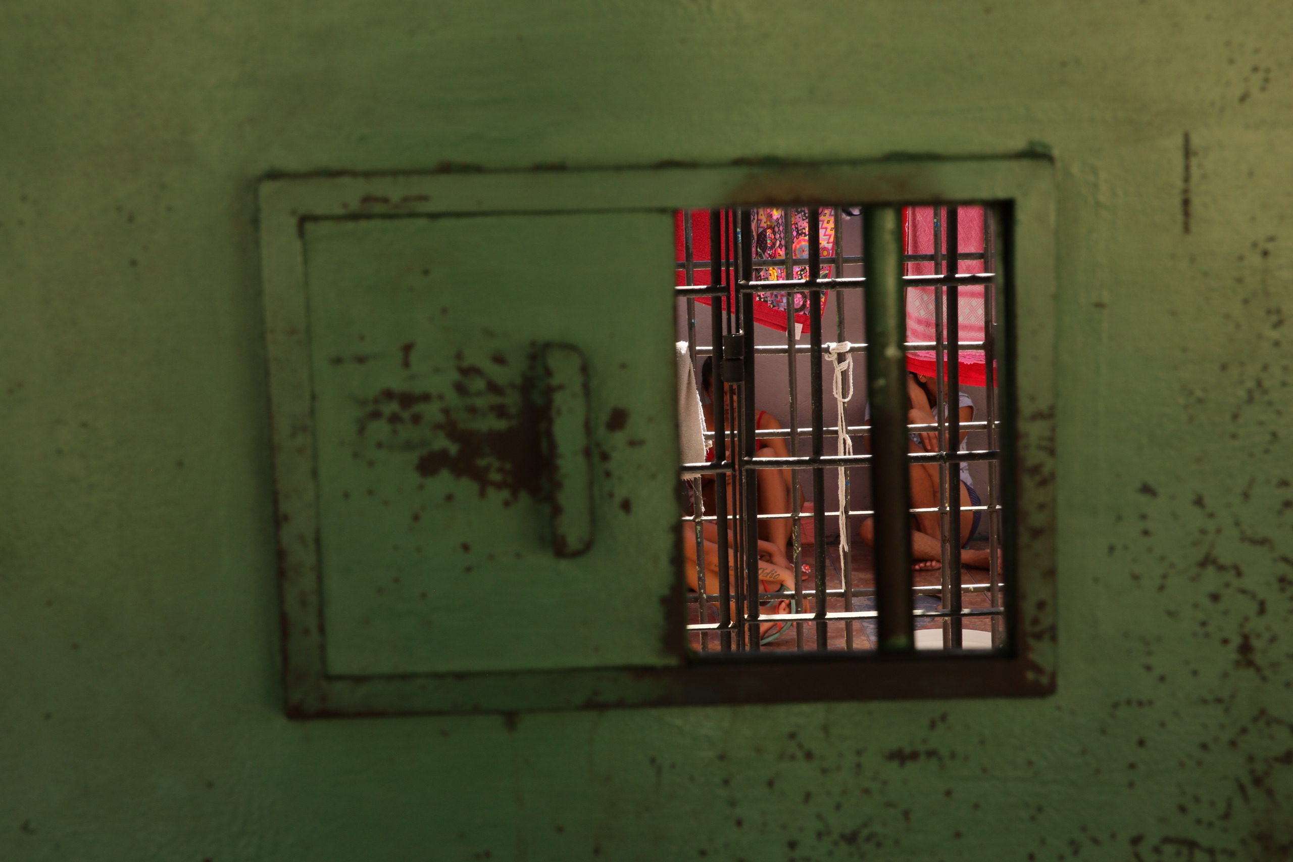 Imagem mostra uma cela de cadeia e atrás mulheres sentadas no chão