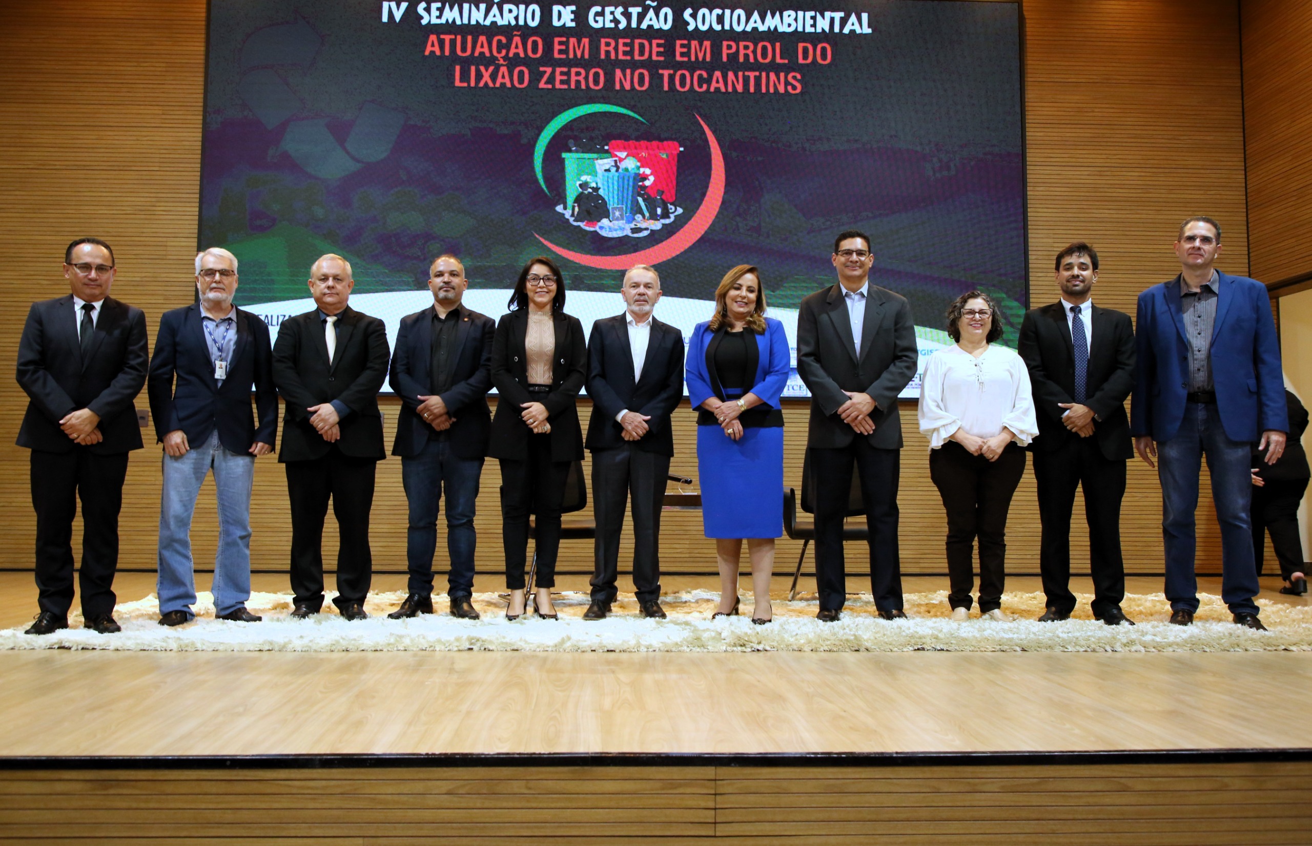 Fotografia colorida onde um grupo de onze pessoas estão posando para foto sobre um palco de madeira clara, e ao fundo está um painel do evento 