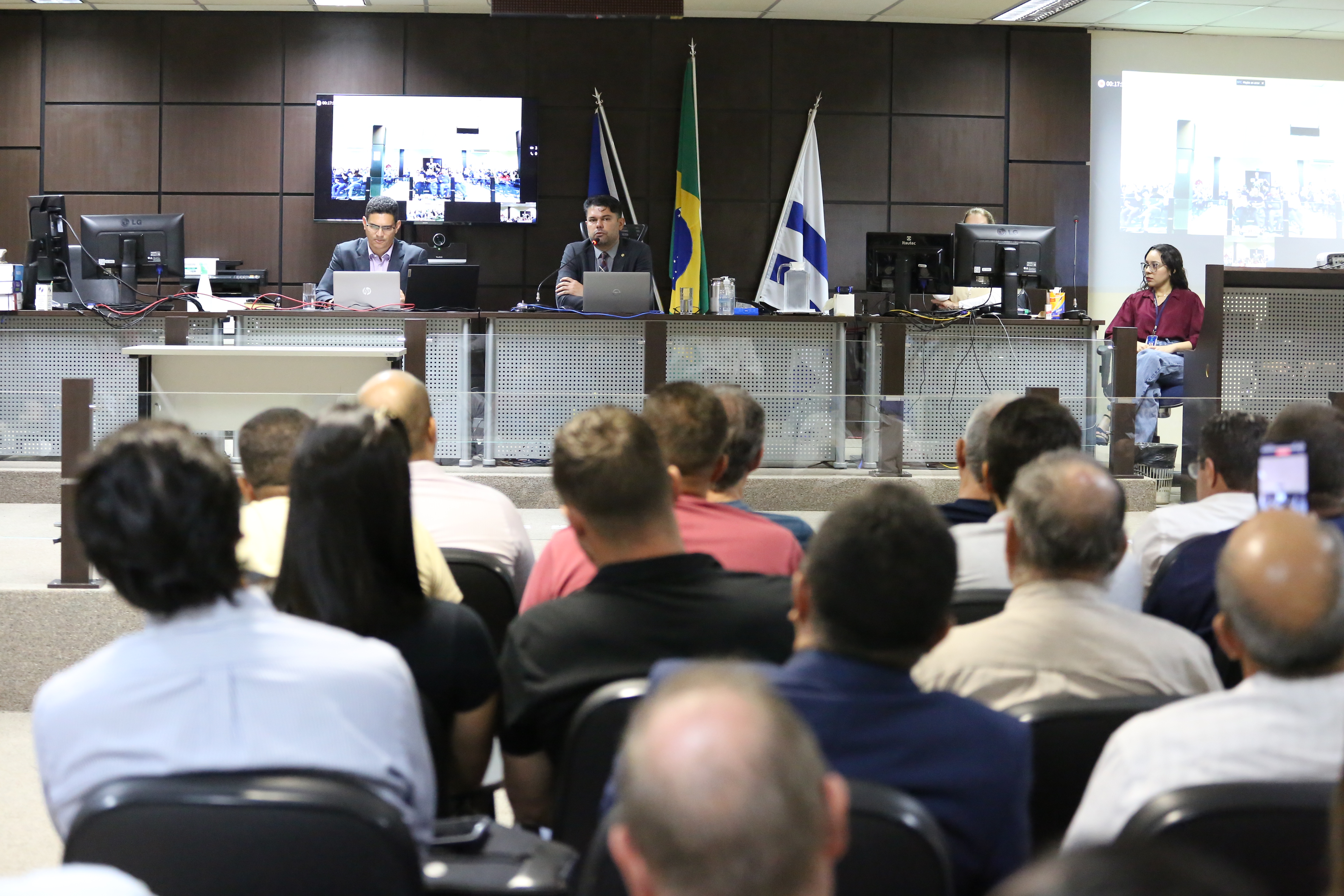 foto em formato paisagem revela pessoas de costas, entadas assistindo um magistrado sentado e fala ao microfone em audiencia publica realizada no salão do juri.