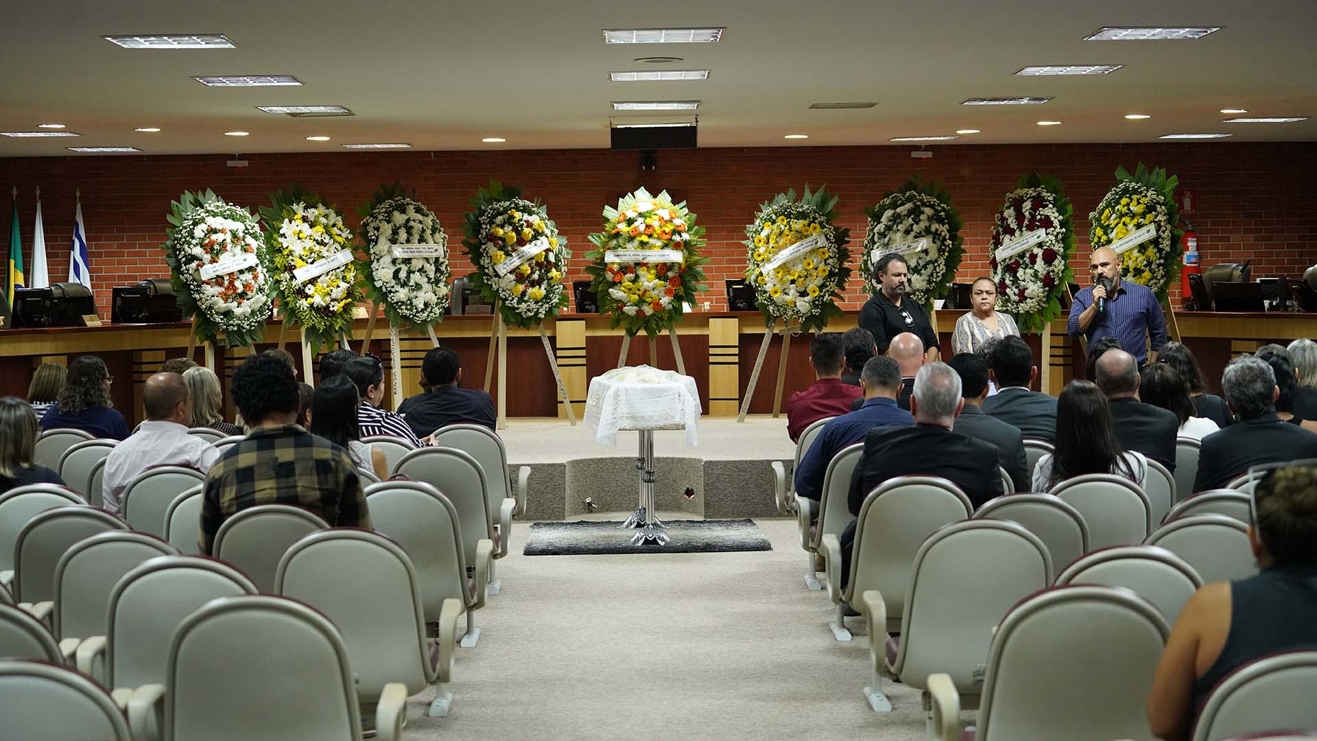 Fotografia colorida que mostra um salão com pessoas sentadas de costas a observar um caixão ao centro; ao fundo há 8 coroa de flores