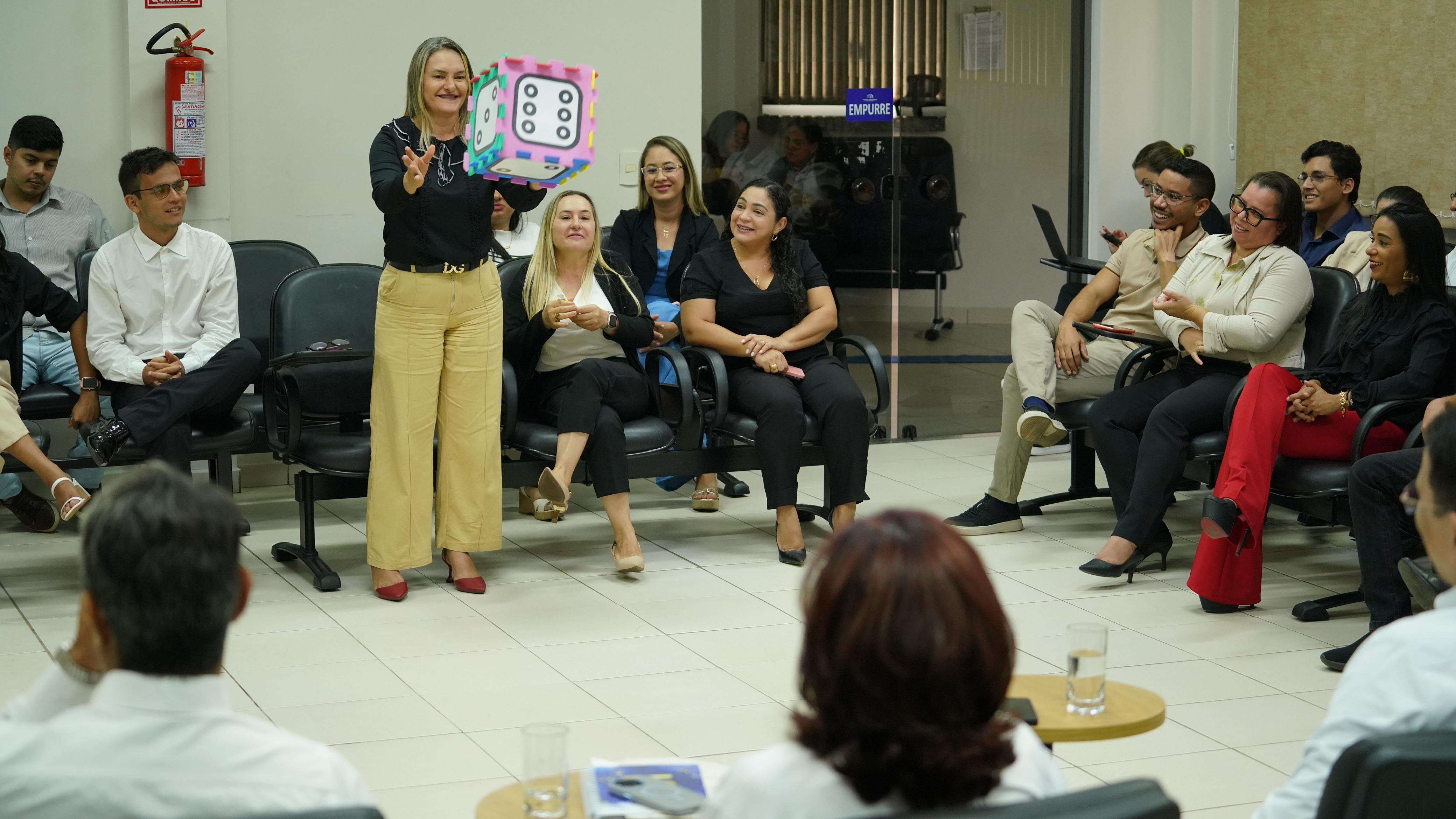 Imagem colorida que mostra uma sala lotada de pessoas sentadas e sorridentes observando uma mulher loira, de calça clara e camisa preta, jogando um dado para o alto