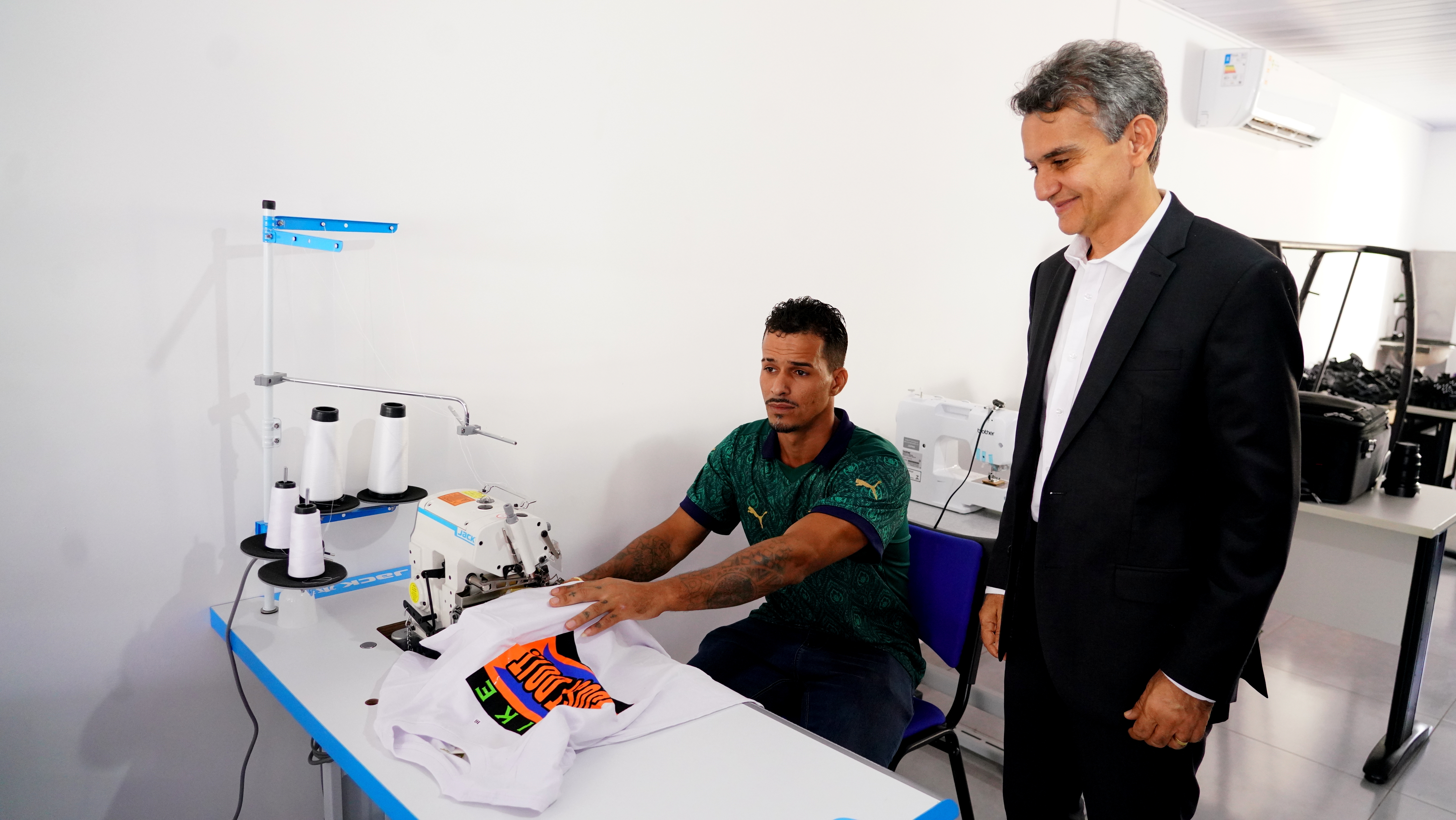 Fotografia colorida que mostra um homem costurando, sentando, e outro homem em pé observando. O homem sentado usa camiseta verde, o homem em pé usa terno e gravata 