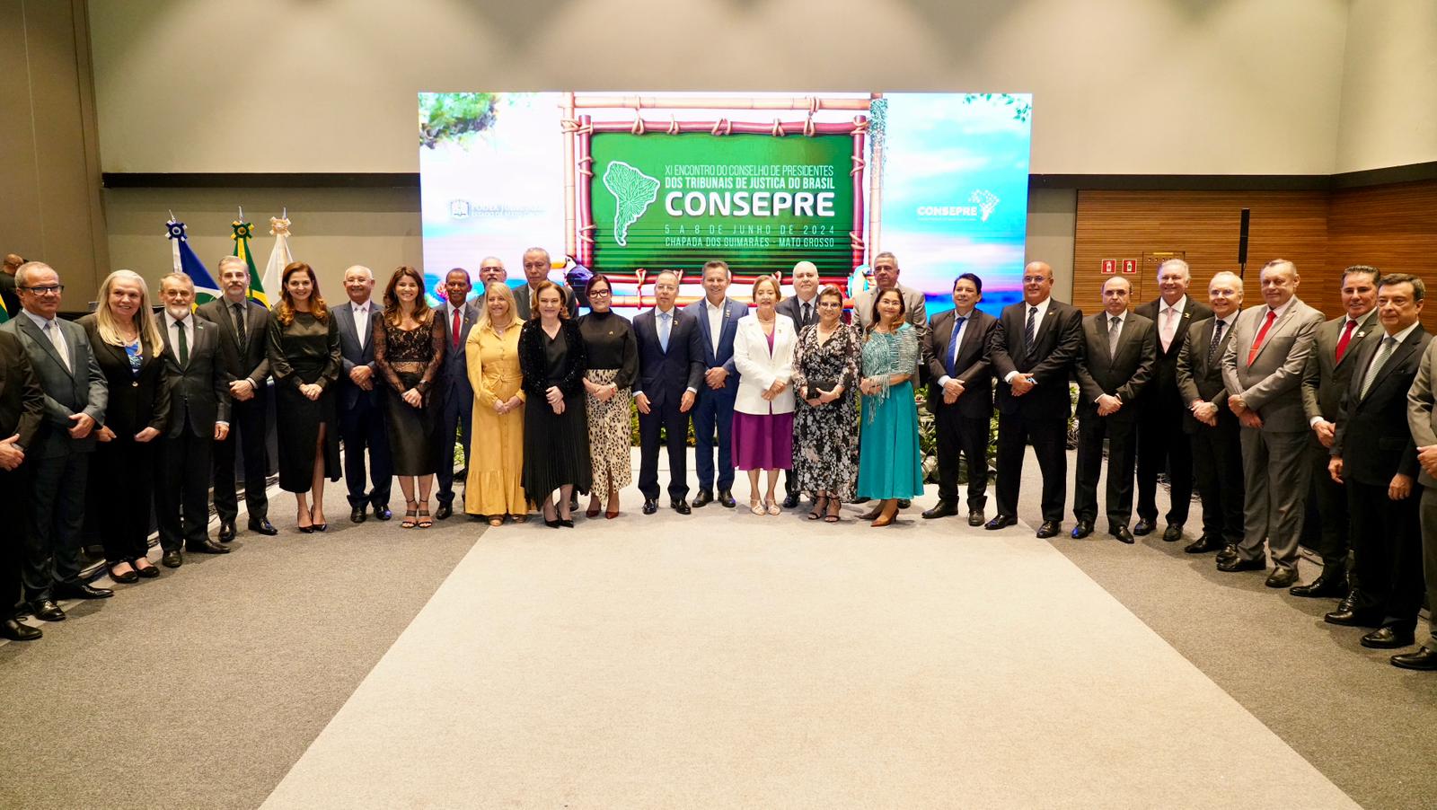Fotografia colorida onde um grupo de magistrados está com um painel da Consepre verde no fundo. Eles estão de pé em um chão claro com um carpete cor de leite no meio 