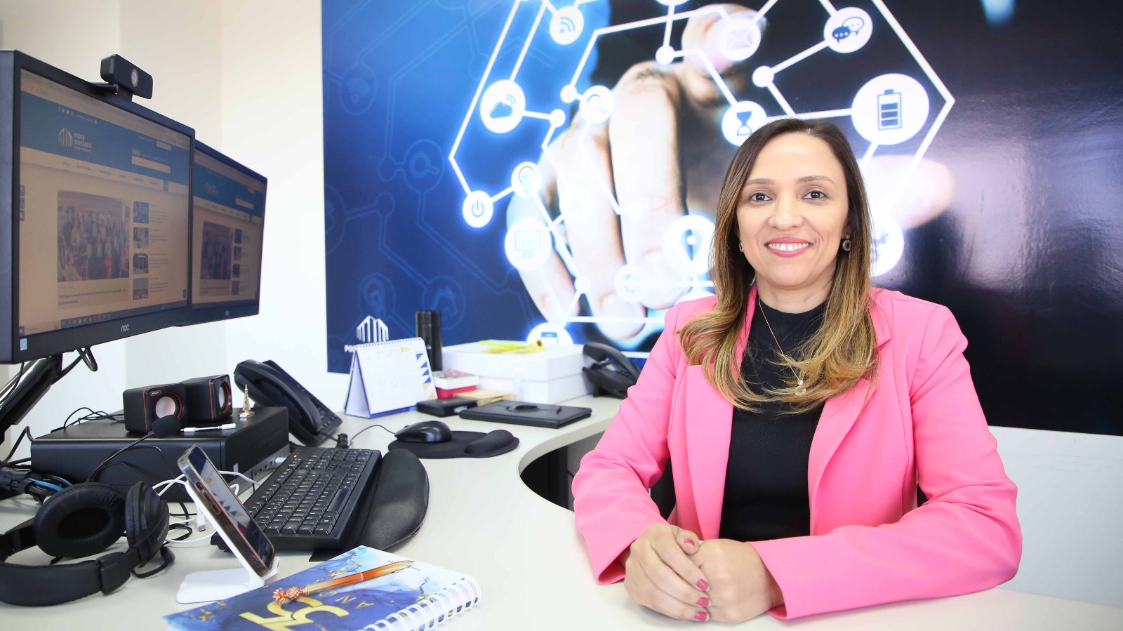 Alice Carla, diretora da Dtinf, sentada, de frente, posando para a foto; ela usa uma blusa preta e um blazer rosa; à esquerda, dois monitores, CPU, teclado, mouse, telefones e outros objetos sobre à mesa; ao fundo uma imagem que remete à tecnologia