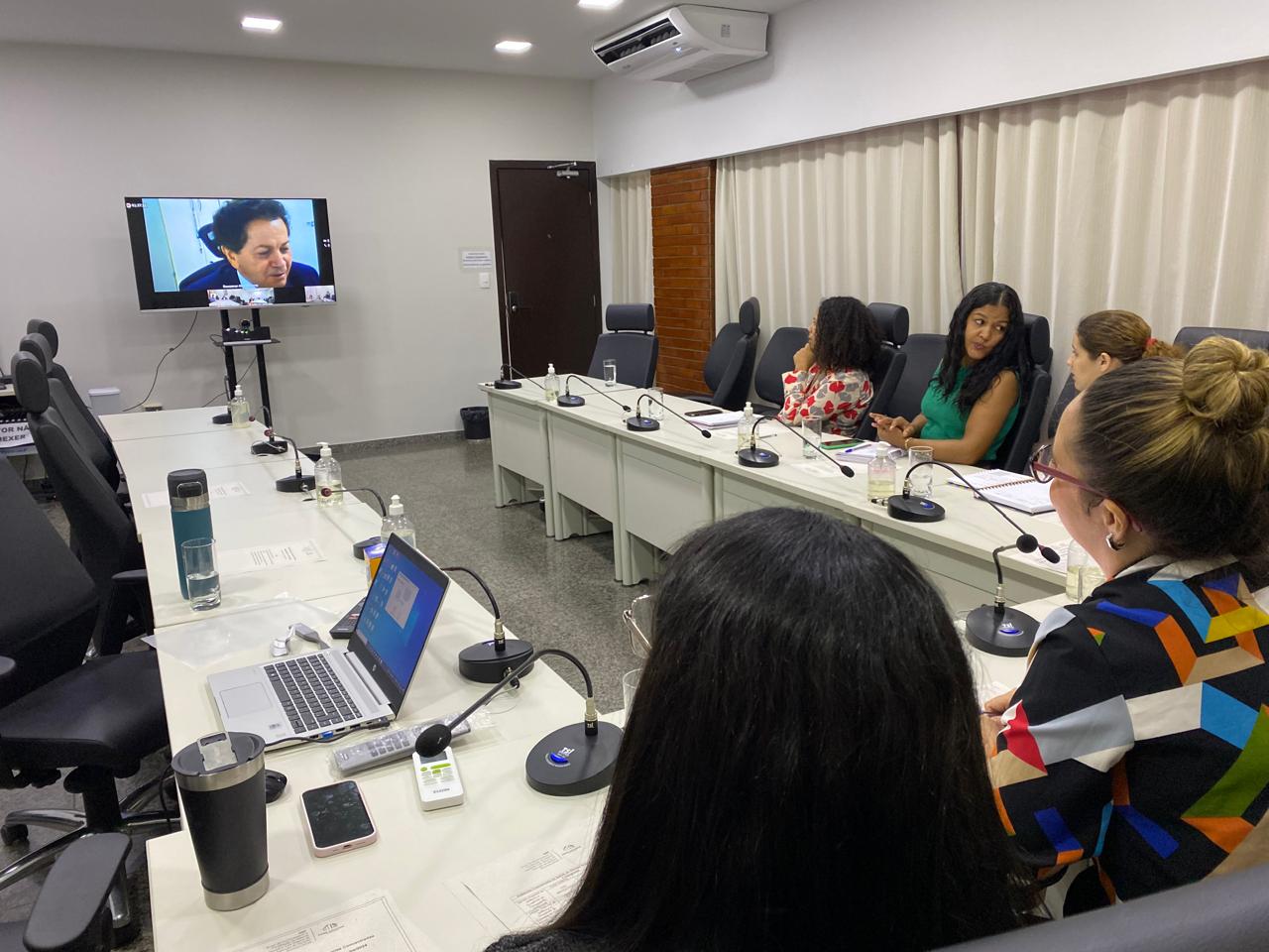 Vários técnicos reunidos em uma sala, sentados em cadeiras dispostas em mesas brancas; paredes e cortinas brancas; ao fundo um monitor com projetando a imagem do juiz Deusamar