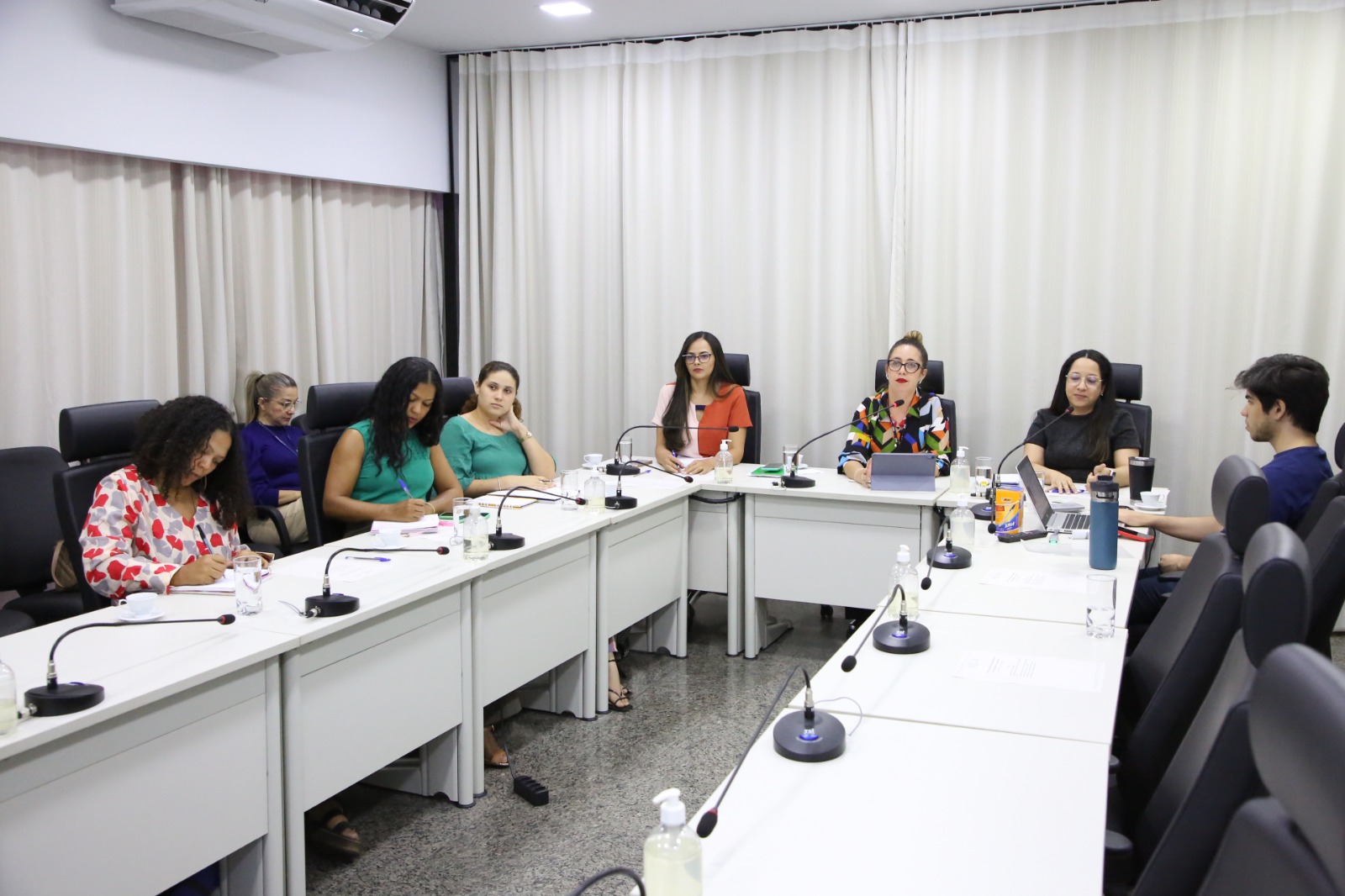 Sala de reunão com mesas brancas dispostas em formato em "U", com servidores e técnicos sentados, reunidos