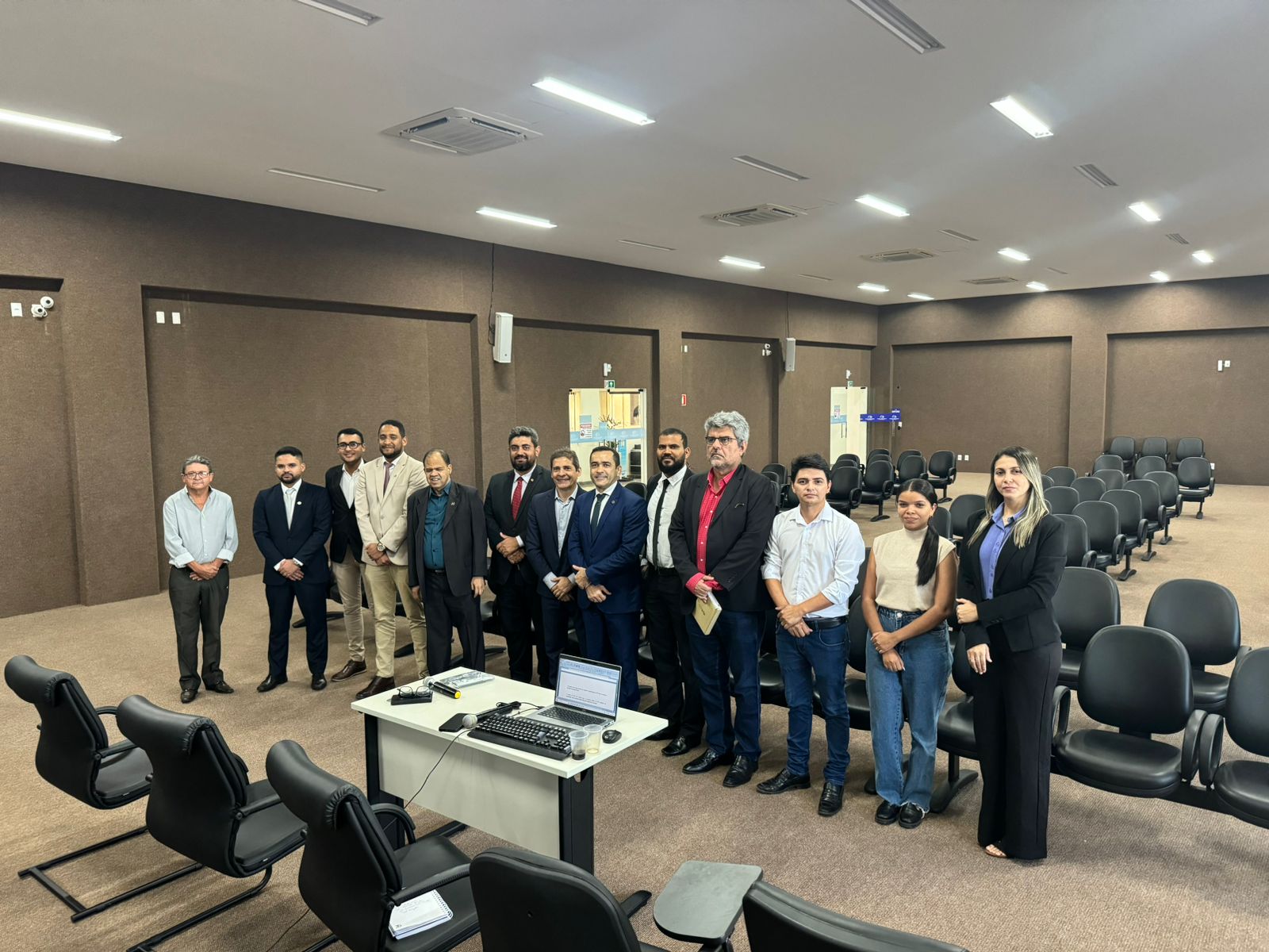 Fotografia colorida onde um grupo de 13 pessoas, duas mulheres e onze homens, estão posicionados em uma sala de reuniões em frente a fileiras de cadeiras escuras com uma mesa de madeira clara na frente, onde estão posicionados um computador e materiais de escrita 