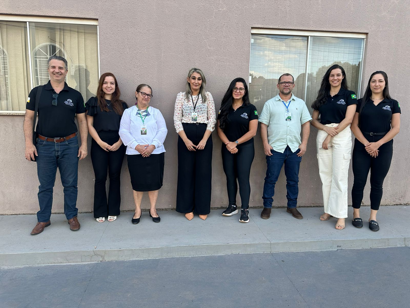 Fotografia colorida onde estão um grupo de seis mulheres e dois homens em pé lado a lado posando para a foto, em frente a uma parede bege com duas janelas de vidro 
