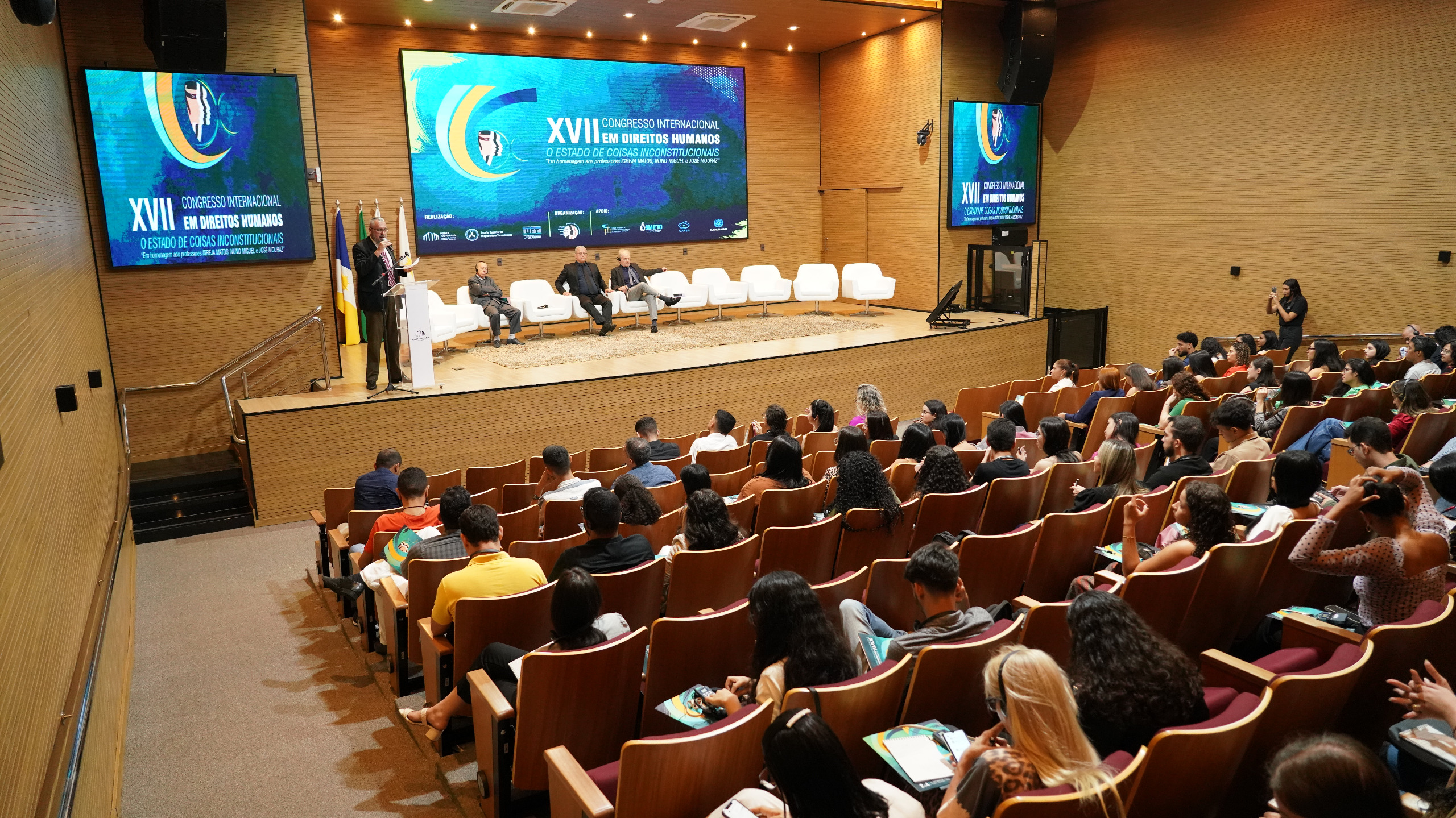 Fotografia colorida que mostra um auditório cheio com um homem falando no palco, enquanto outros três homens o observam.