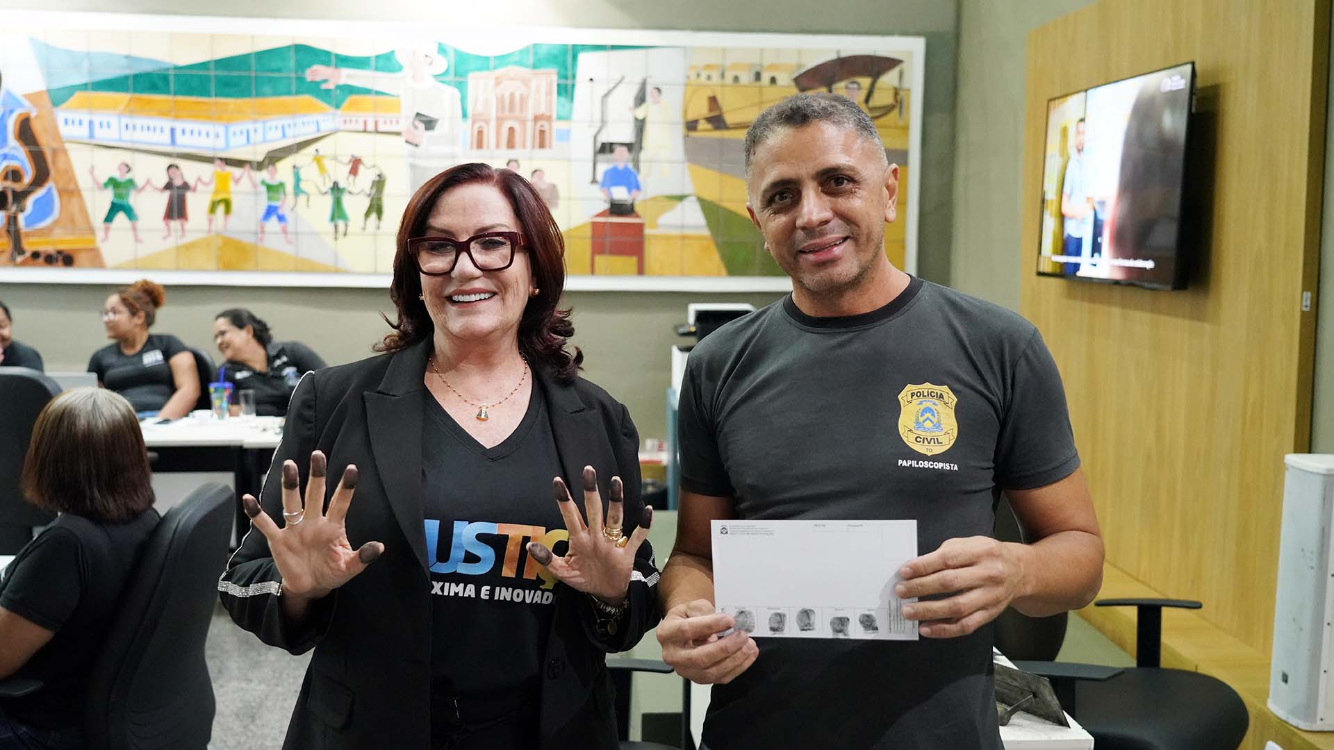 Um homem e uma mulher em pé, com roupas pretas; a mulher é a presidente do TJTO que está mostrando os dedos com as digitais