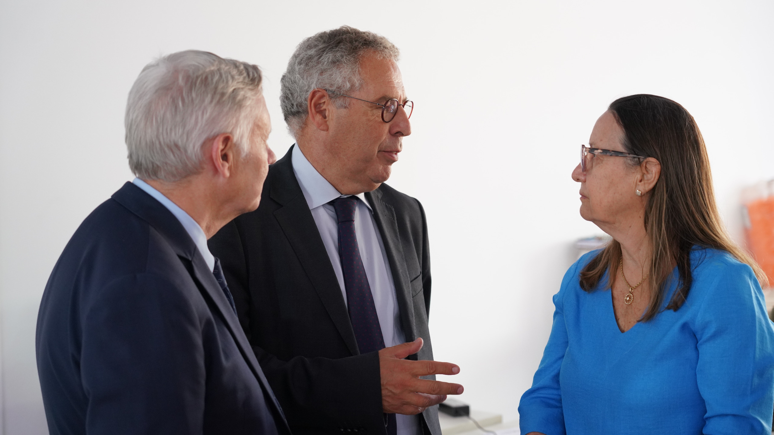 Fotografia colorida que mostra dois homens e uma mulher branca, que usa óculos, conversando