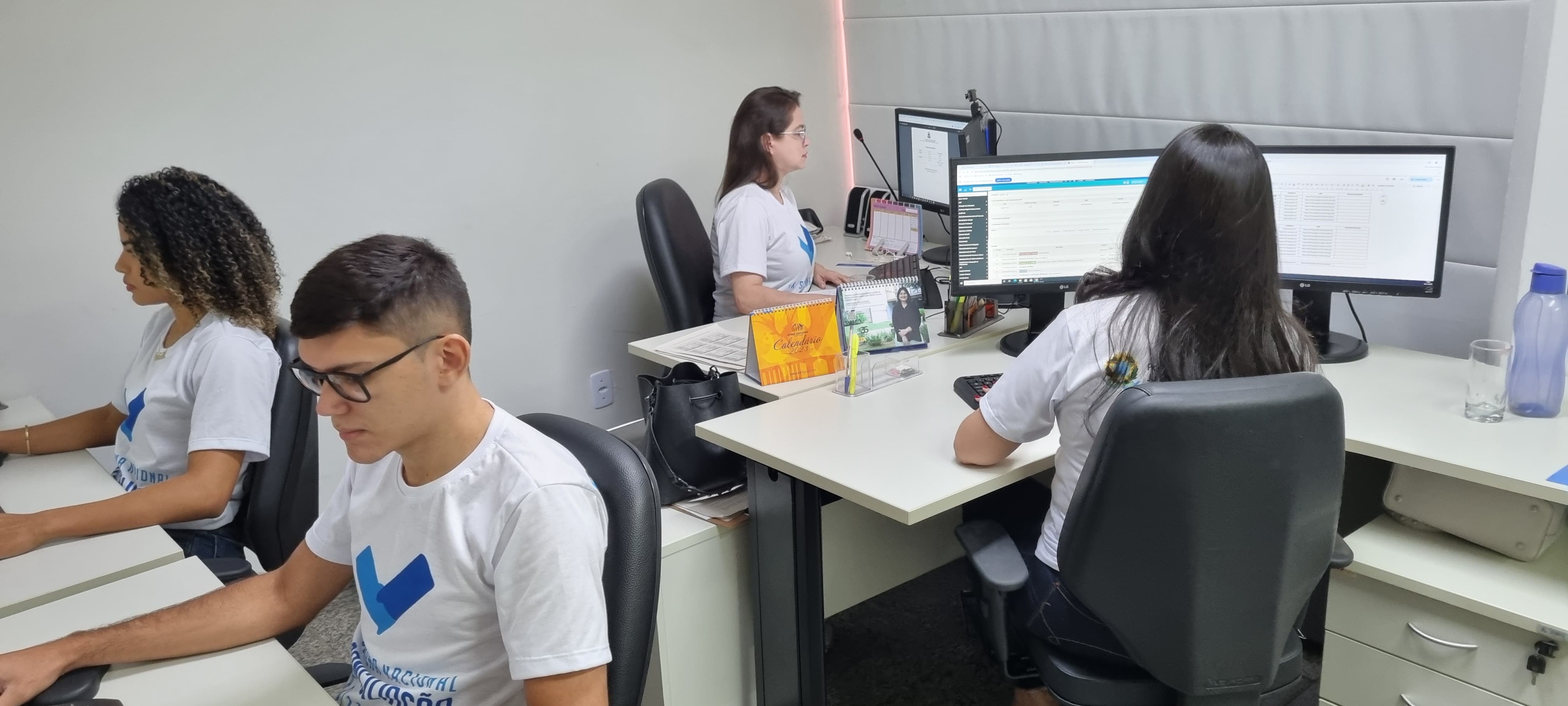 Foto mostra quatro pessoas sentadas em frente à computadores; todos estão de blusa branca