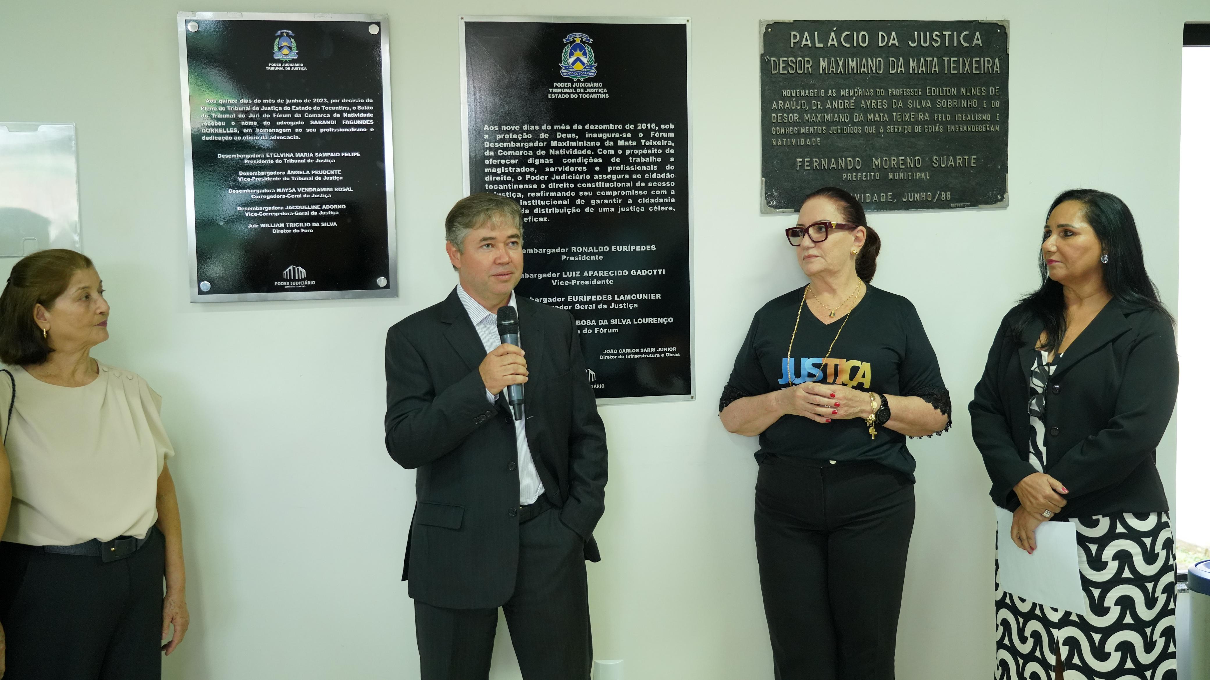 Homem de terno escuro segura um microfone, ao lado dele outras três mulheres em pé escutam  o que ele diz