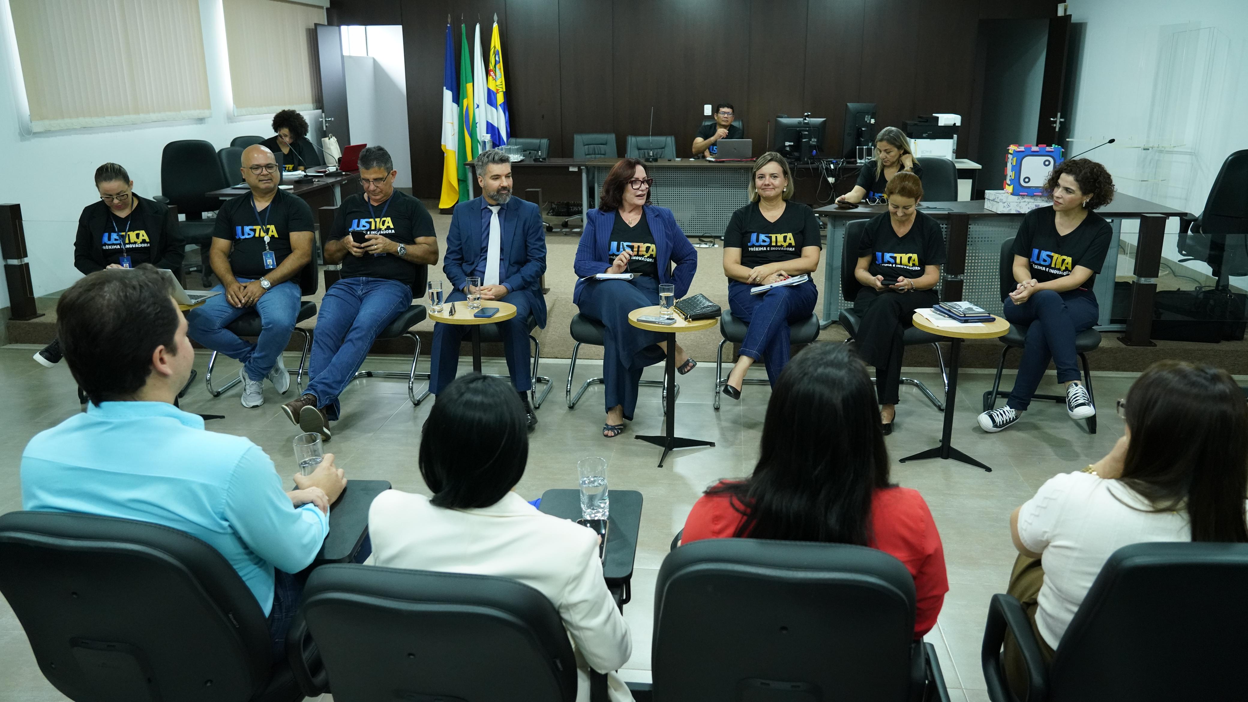 Fotografia colorida que mostra mulheres e homens sentados em cadeiras, dentro do Fórum, conversando. 