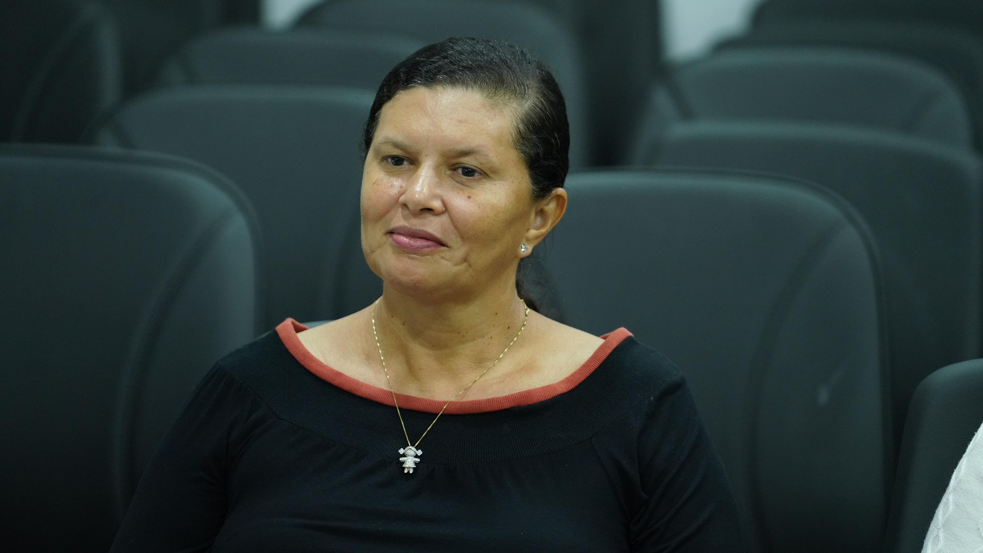 Imagem colorida que mostra uma mulher morena, usando preto, ela está de cabelos presos. 