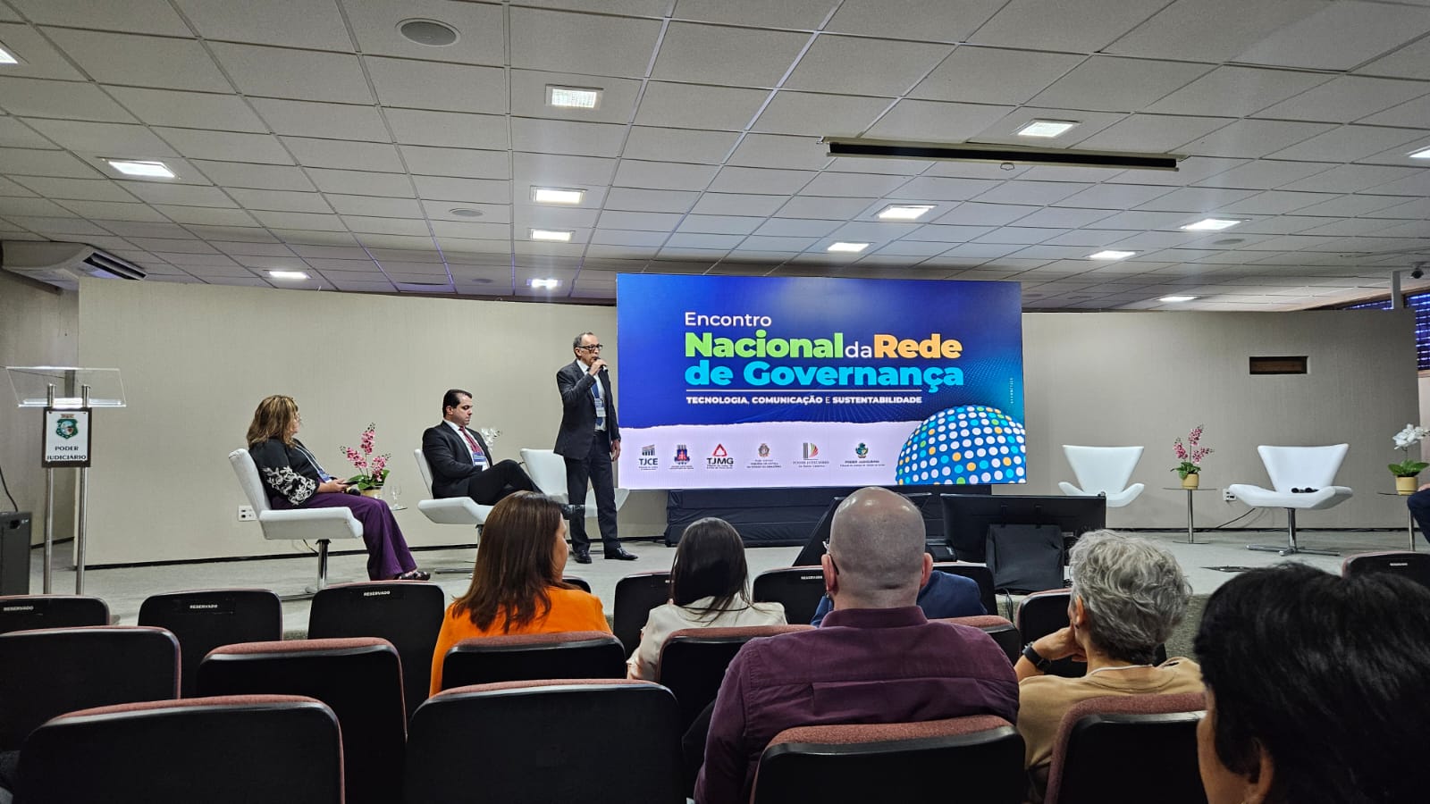 Foto mostra um lugar grande com paredes claras e cadeiras vermelhas; ao palco um homem em pé outro sentado e uma mulher sentada; ao fundo um telão