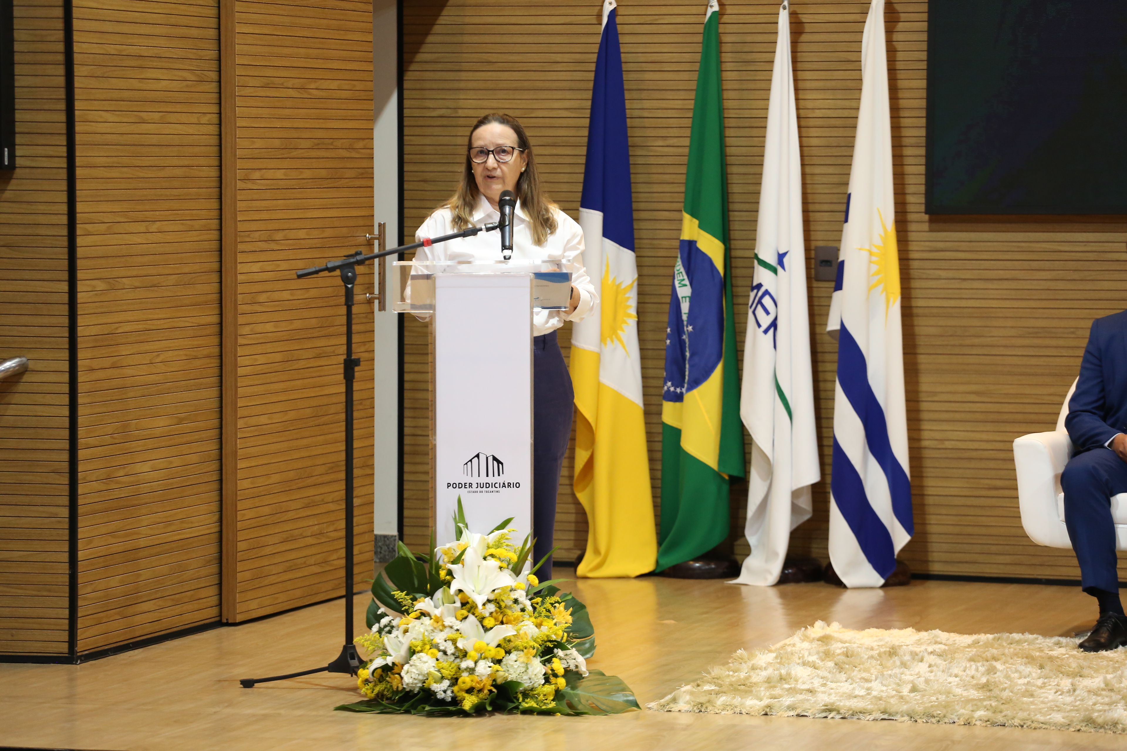 Fotografia colorida que mostra da desa. Maysa Vendramini, uma mulher branca de óculos e cabelos castanhos claros, usando blusa branca e calça preta, ela fala ao microfone