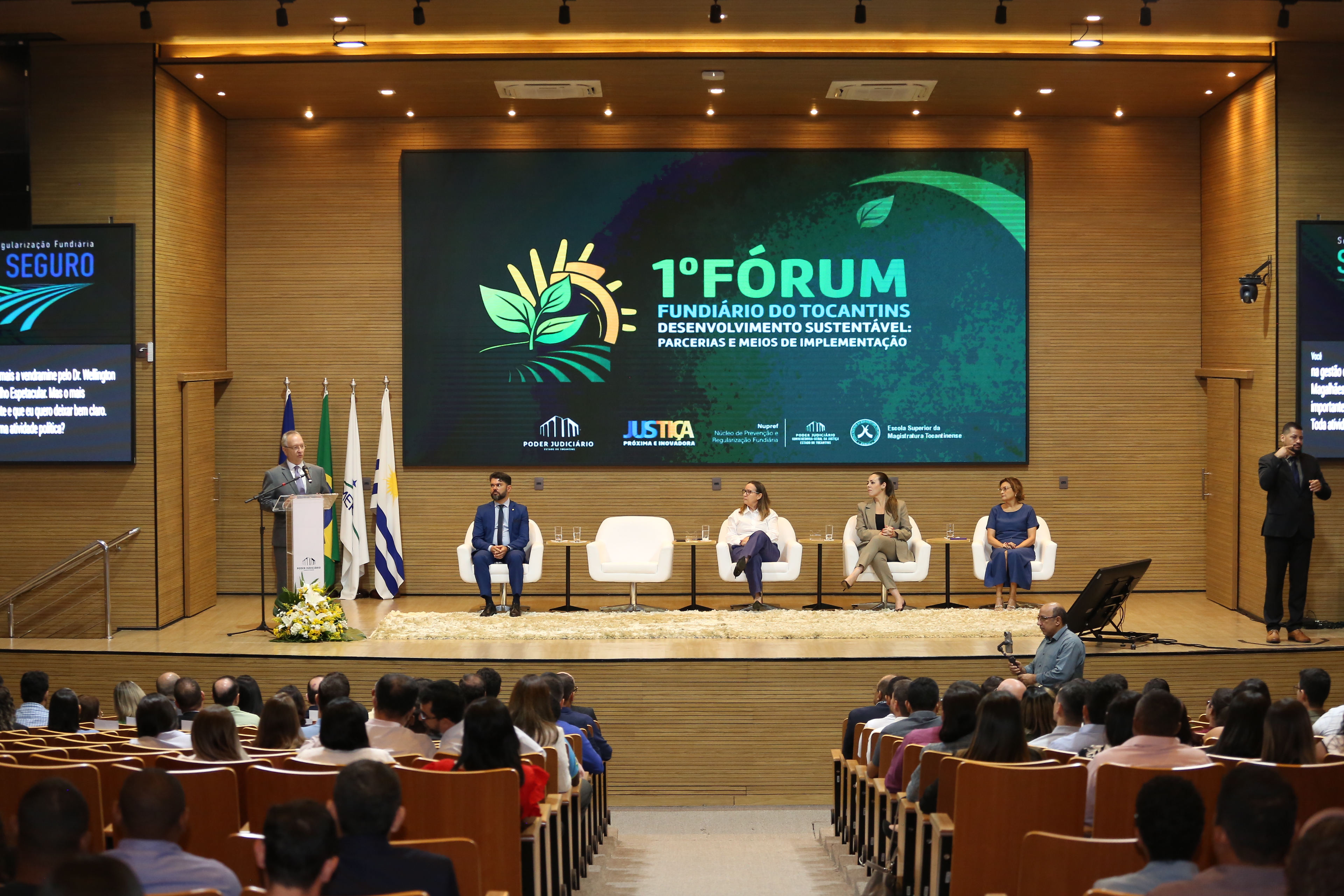 Fotografia colorida que mostra o des. Pedro Nelson, um homem branco, cabelos grisalhos, que está usando terno cinza, falando ao público do evento.