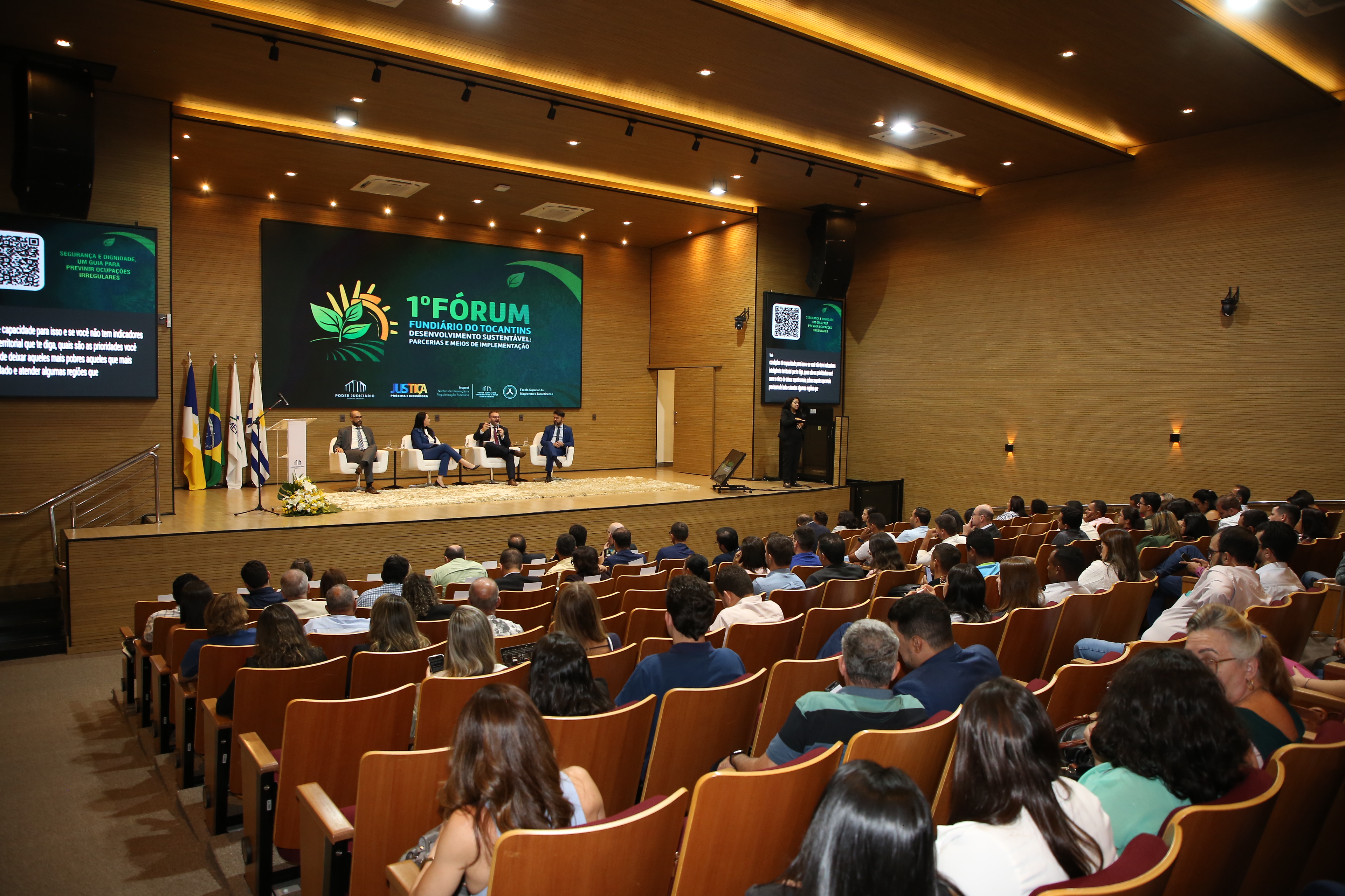 Fotografia colorida que mostra autoridades em um palco com fundo do 1º Fórum Fundiário do Tocantins e um auditório lotado de pessoas sentadas 