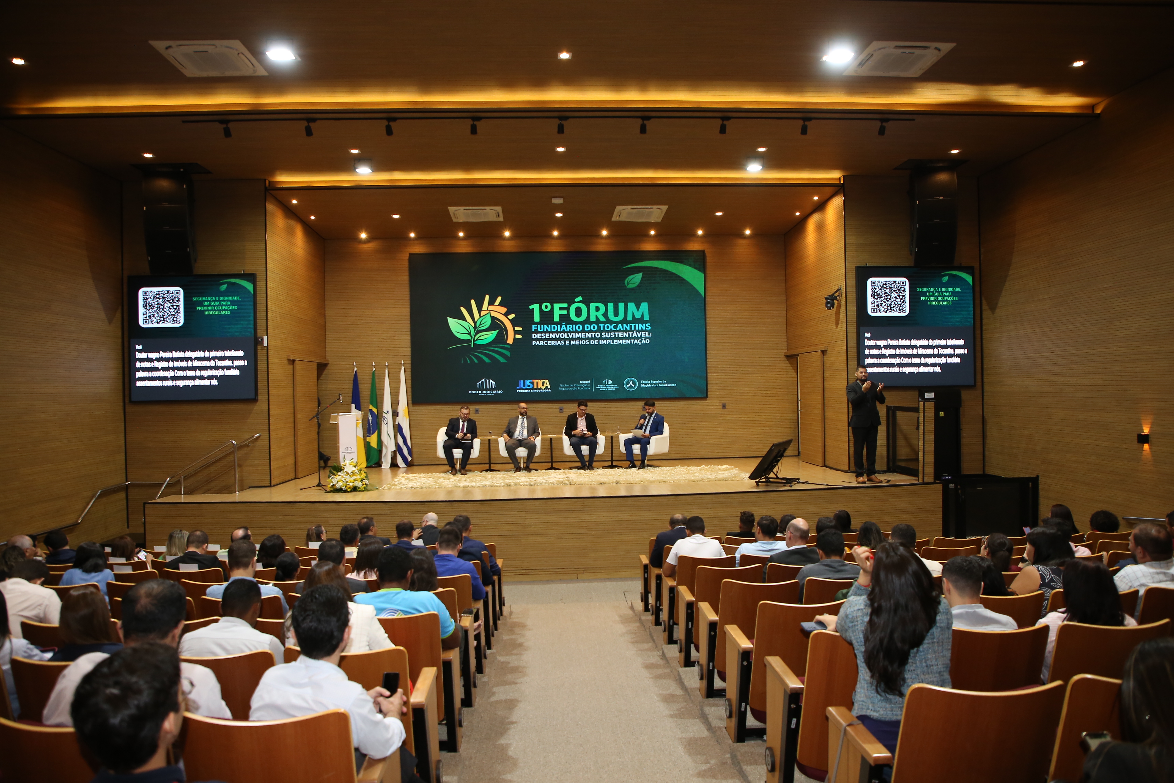 Foto colorida horizontal mostra o auditório do TJTO com pessoas sentadas na plateia assistindo no palco um debate entre quatro homens sentados falando ao microfone.