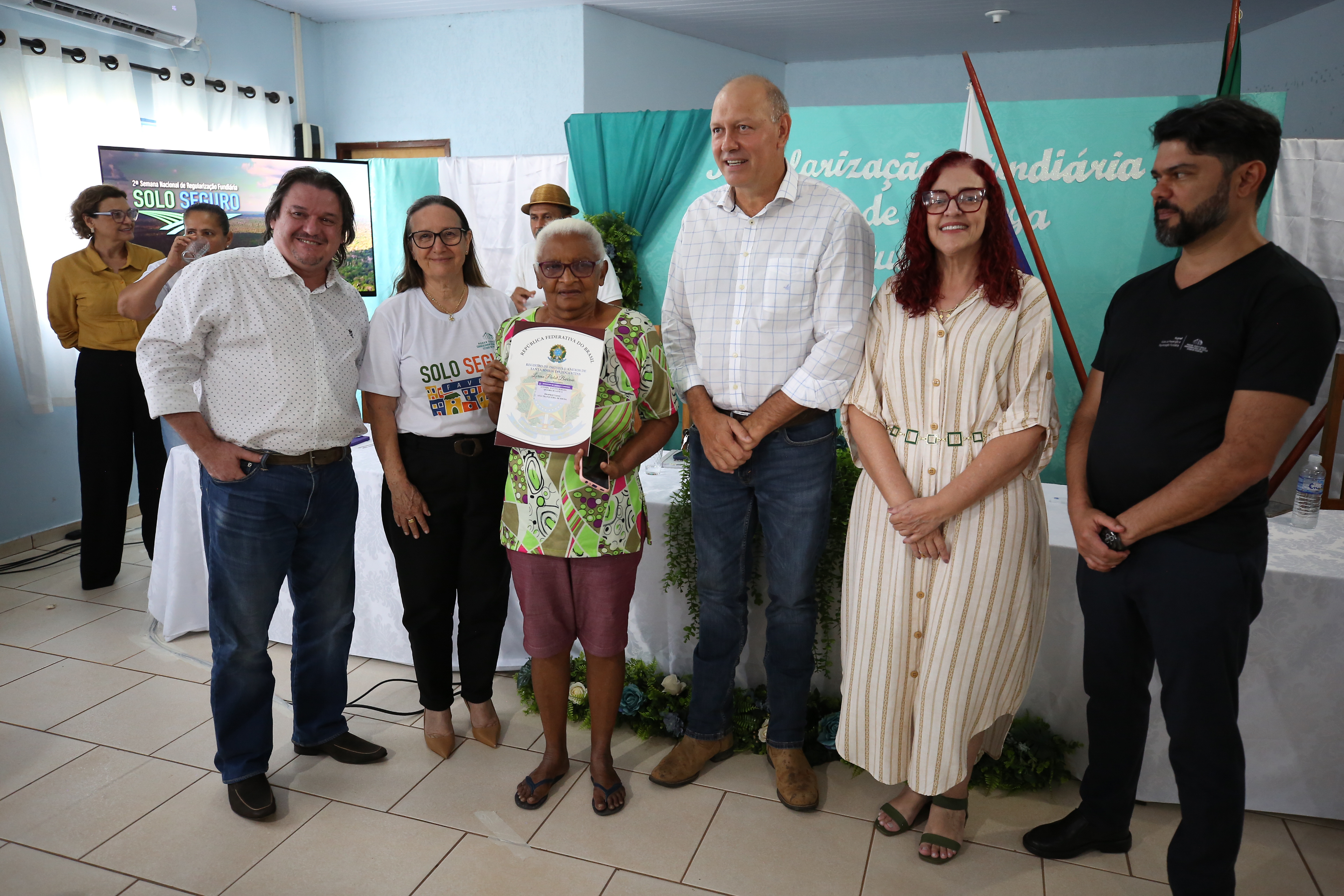 Fotografia colorida que mostra uma mulher negra segurando um documento de posse ao lado de um homem moreno de barba e de uma mulher branca