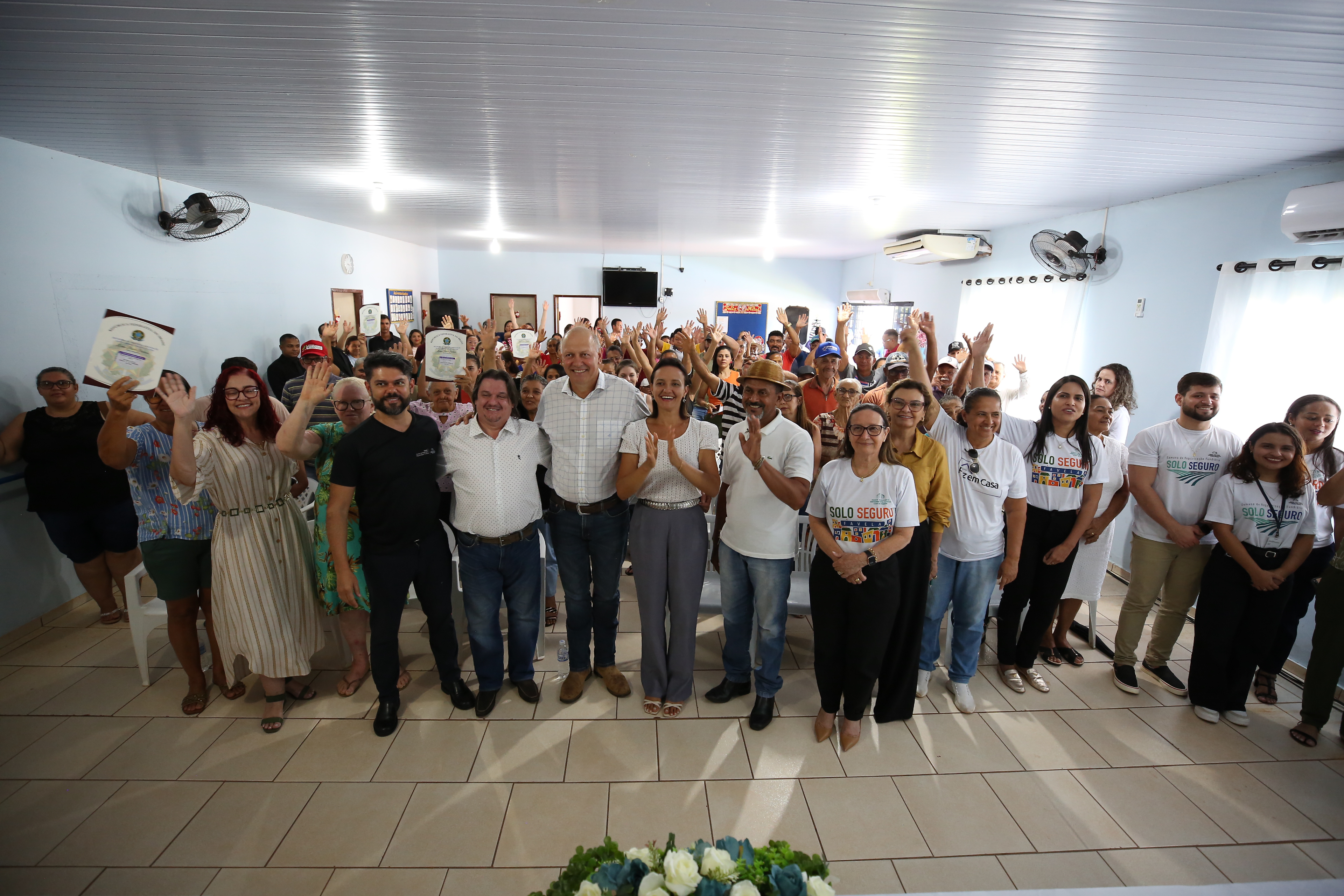 Fotografia colorida que mostra várias pessoas em pé aplaudindo, eles estão com documentos nas mãos posam pra foto