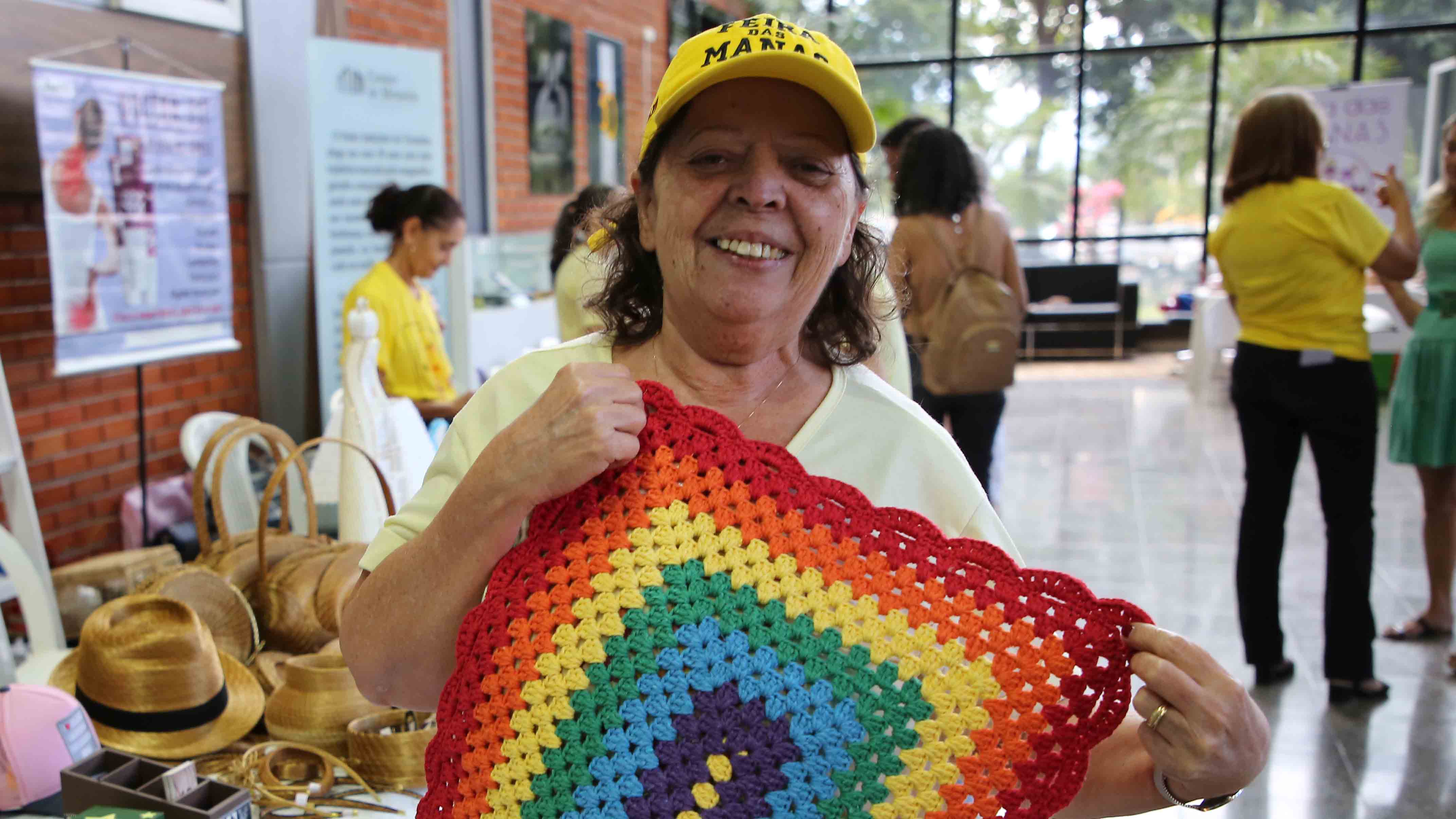 Foto colorida de uma mulher segurando um tapete de crochê, ela sorri. Ao fundo mulheres vendem na feira ecosolidária